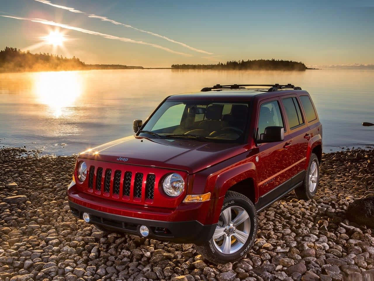 Jeep Patriot 桌布 壁紙 — 一輛亮紅色的Jeep Patriot停泊在岩石湖岸邊，太陽升起，金色光芒灑在霧氣瀰漫的水面上。
