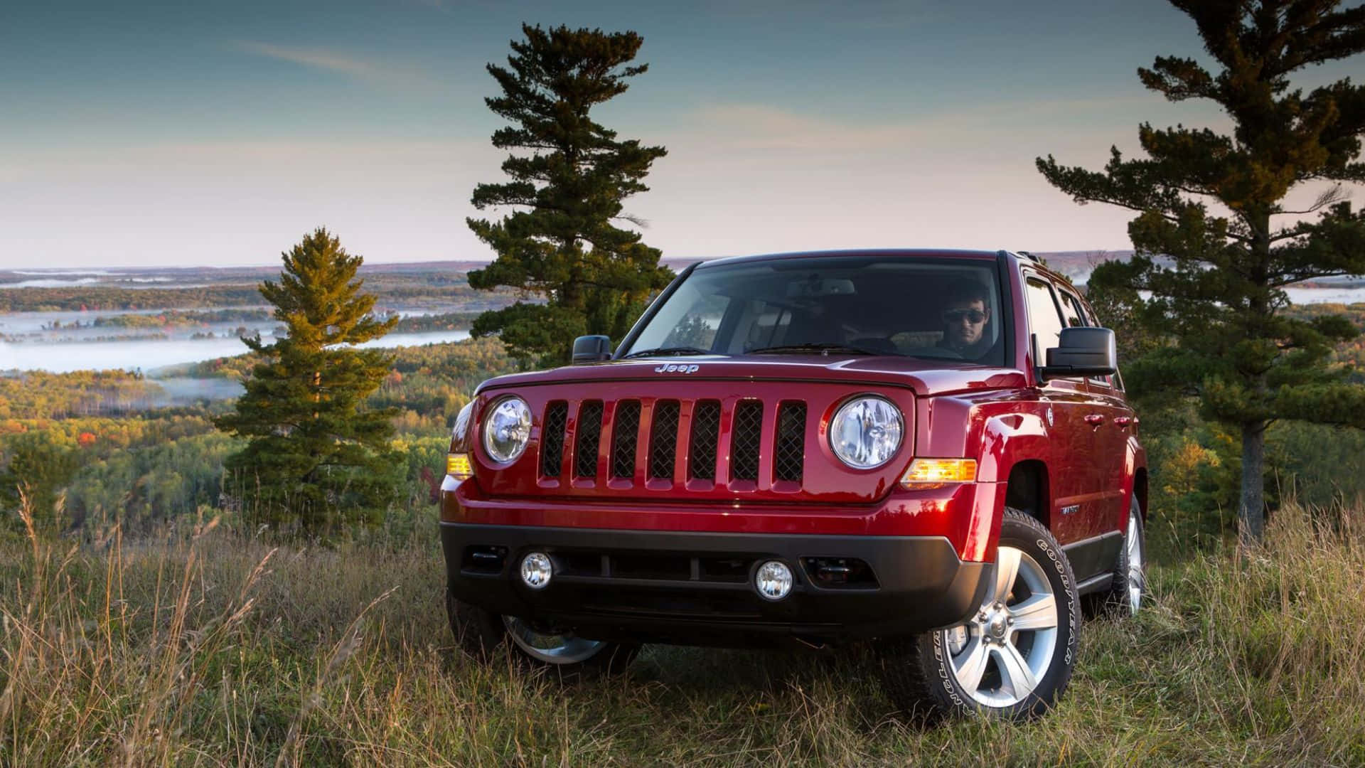 Jeep Patriot 桌布 壁紙 — 一輛亮紅色的Jeep Patriot停泊在長滿青草的山坡上，俯瞰著被秋樹環繞的霧氣瀰漫的湖泊。