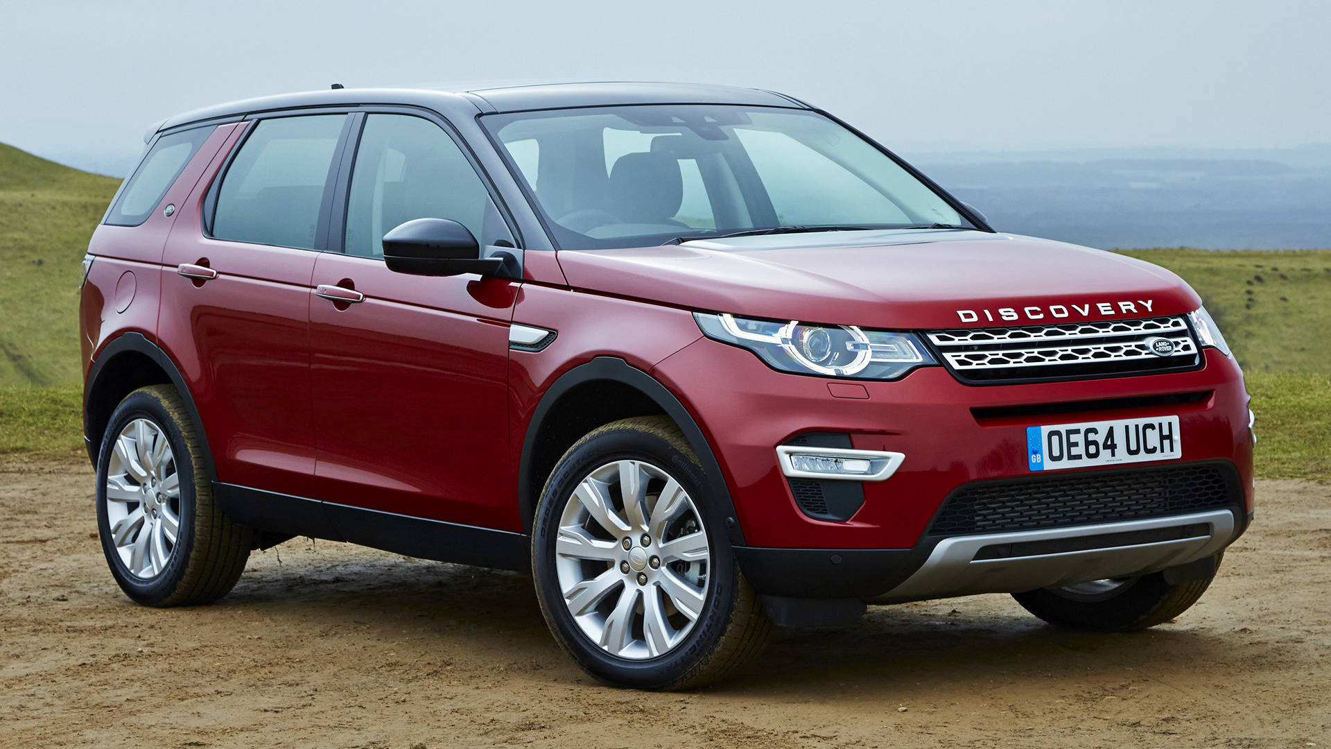 land rover iPhone 桌布 壁紙 — 一輛亮紅色的Land Rover Discovery SUV停在一條泥土路上，背景是起伏的綠色山丘，天空多雲。