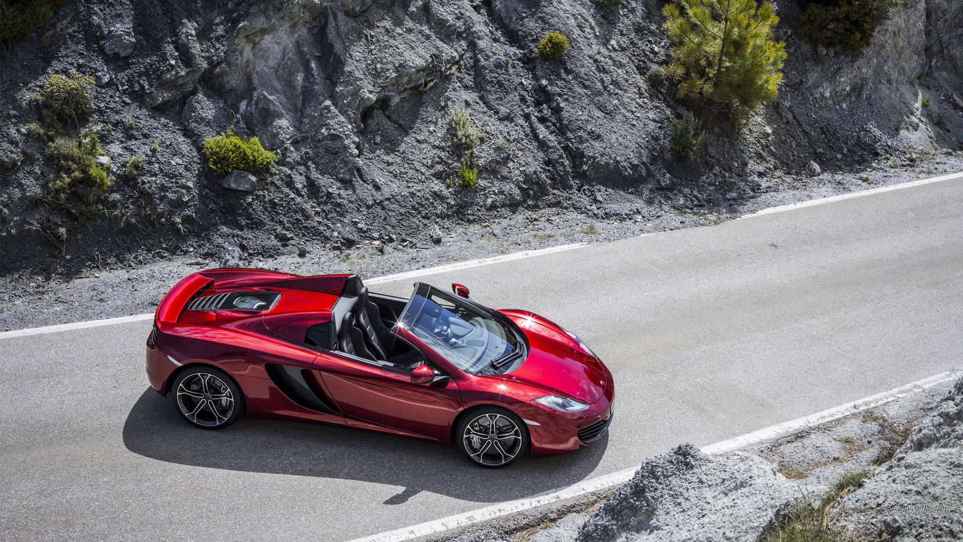 跑車 桌布 壁紙 — 一輛亮紅色的McLaren跑車在明亮的日光下行駛在一條蜿蜒的山路上，兩旁是崎嶇的灰色懸崖和稀疏的綠植。