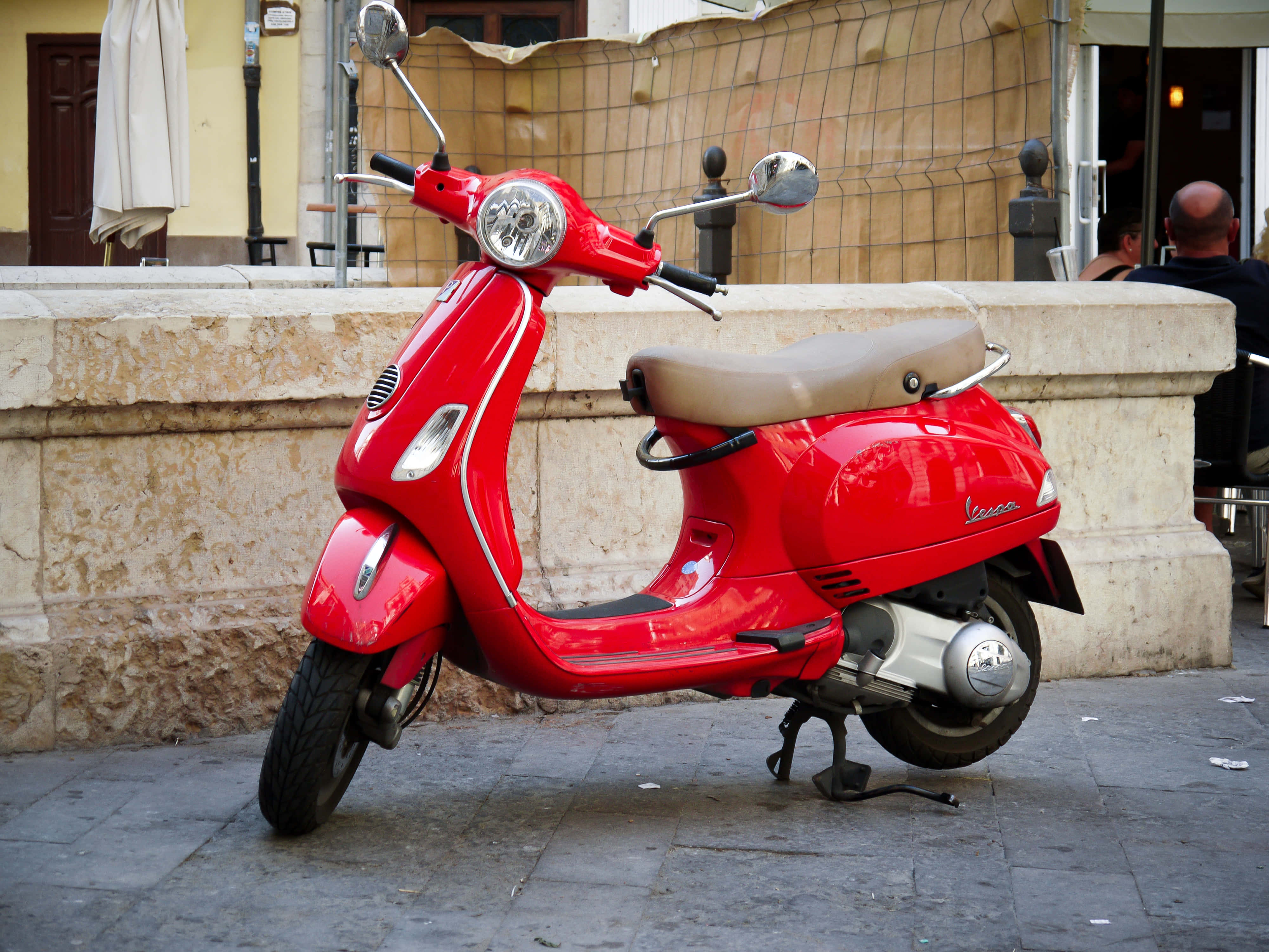 scooter 桌布 壁紙 — 一輛亮紅色的Vespa摩托車停在石頭人行道上，旁邊是米色石牆，背景有人。