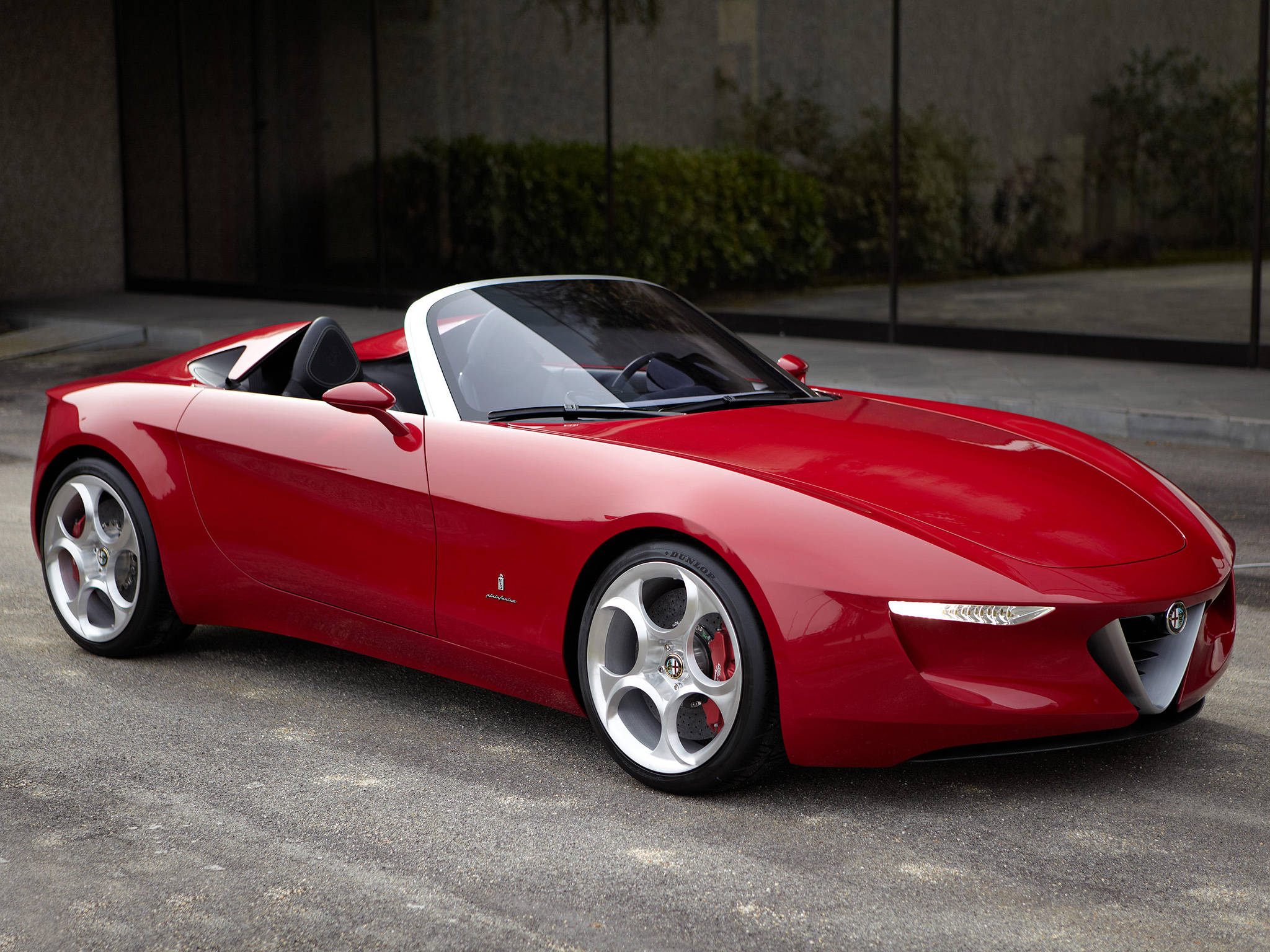 alfa romeo iPhone 桌布 壁紙 — 一輛亮紅色的愛快羅密歐Spider敞篷車停在柏油路上，背景有現代玻璃建築和綠植。