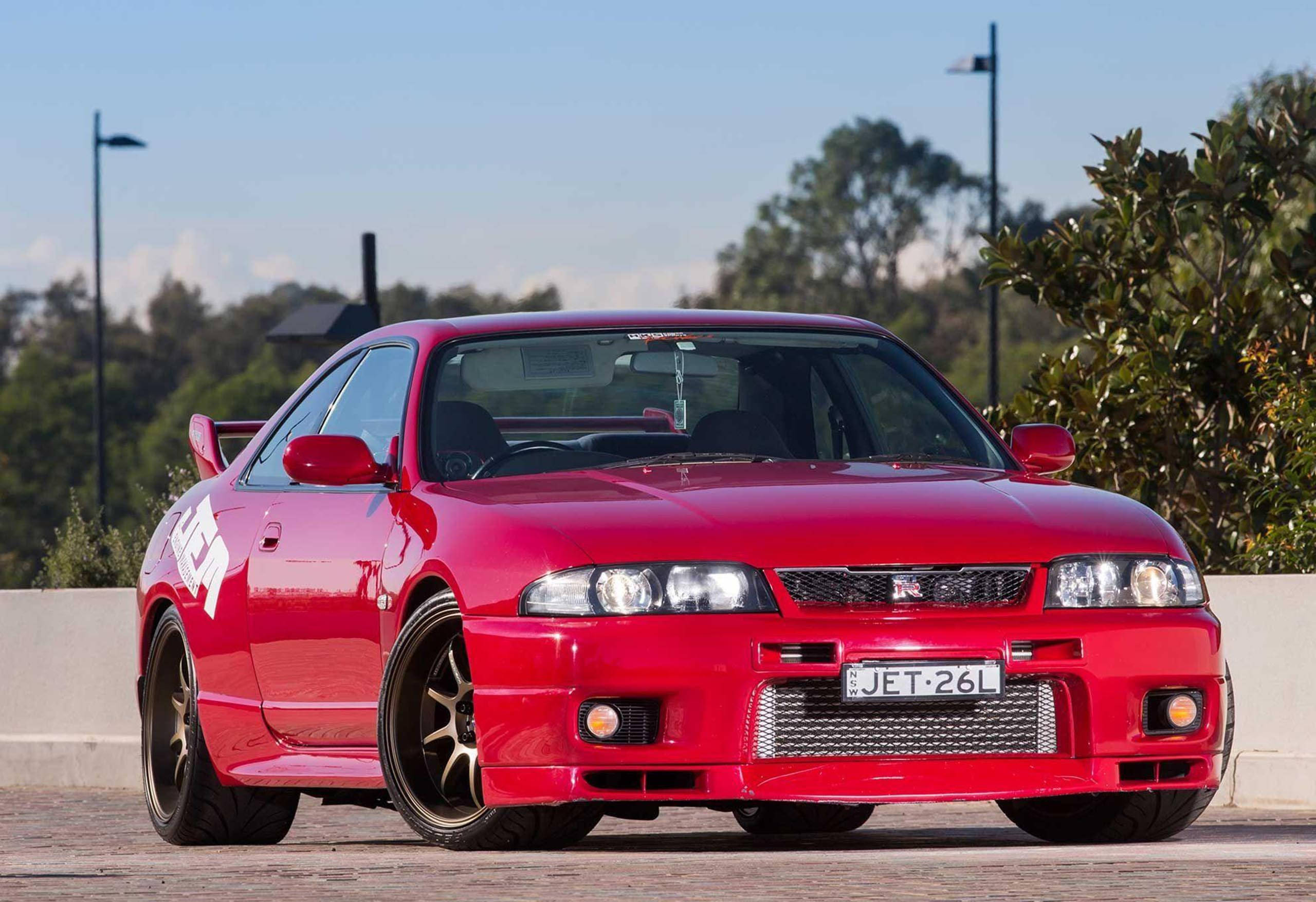 Gtr 桌布 壁紙 — 一輛亮紅色的日產Skyline R34跑車停放在鋪砌區域，周圍有樹木和路燈，天空湛藍。