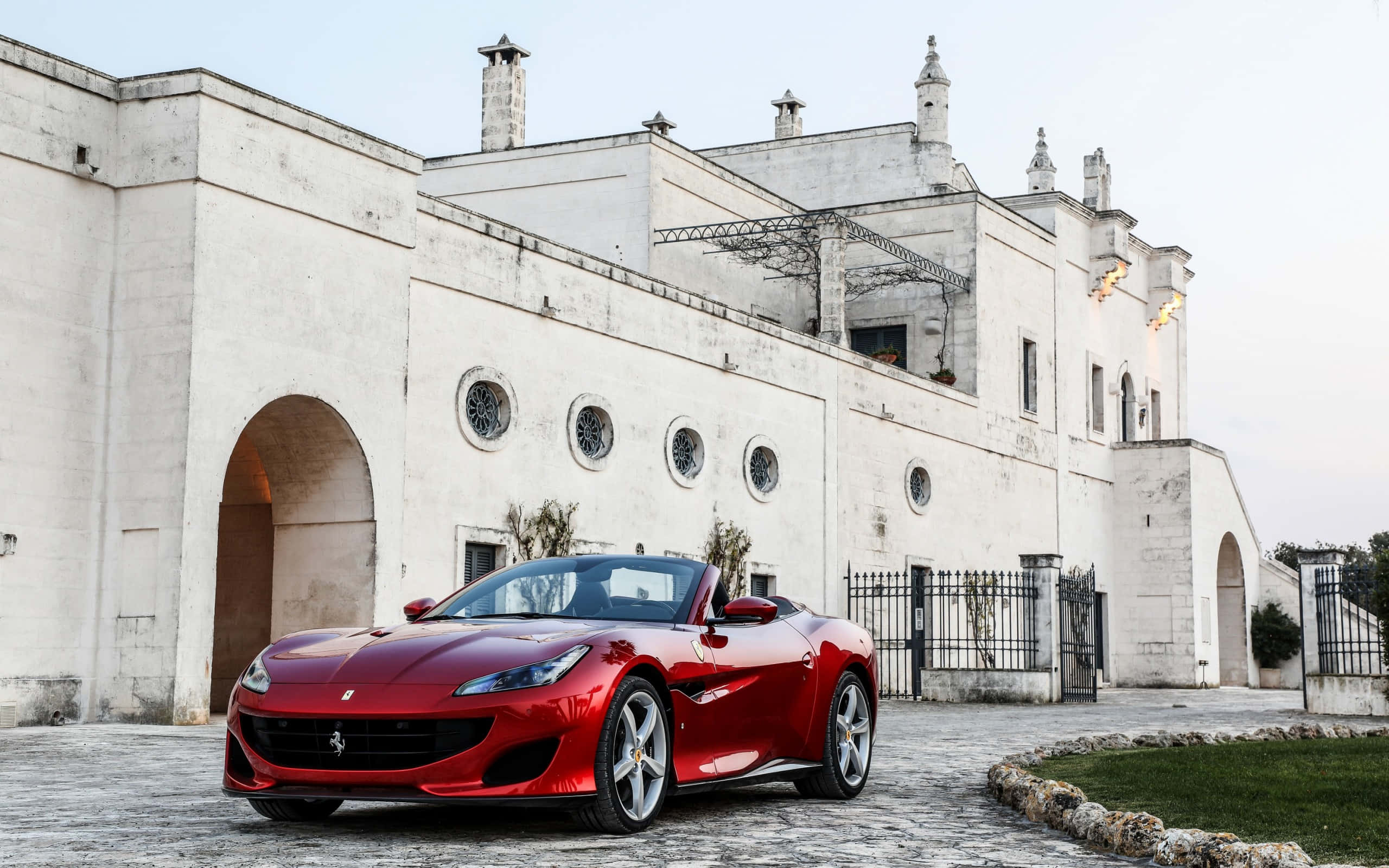 Ferrari Portofino 桌布 壁紙 — 一輛亮紅色的法拉利 Spider 停在鵝卵石路上，前方是一座帶有拱形門廊和圓形窗戶的白色大型歷史建築。