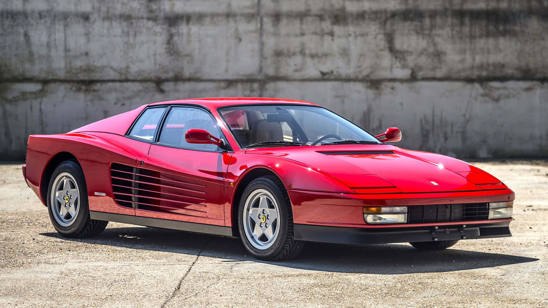 Ferrari Testarossa 桌布 壁紙 — 一輛亮紅色的法拉利 Testarossa 停放在裂開的混凝土上，背景是風化的混凝土牆，在明亮的日光下。