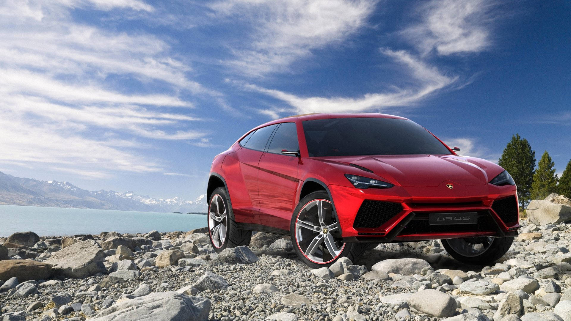 lambo 桌布 壁紙 — 一輛亮紅色的藍寶堅尼 Urus SUV 大膽地停在岩石湖岸上，背景是白雪覆蓋的山脈和明亮的藍天。