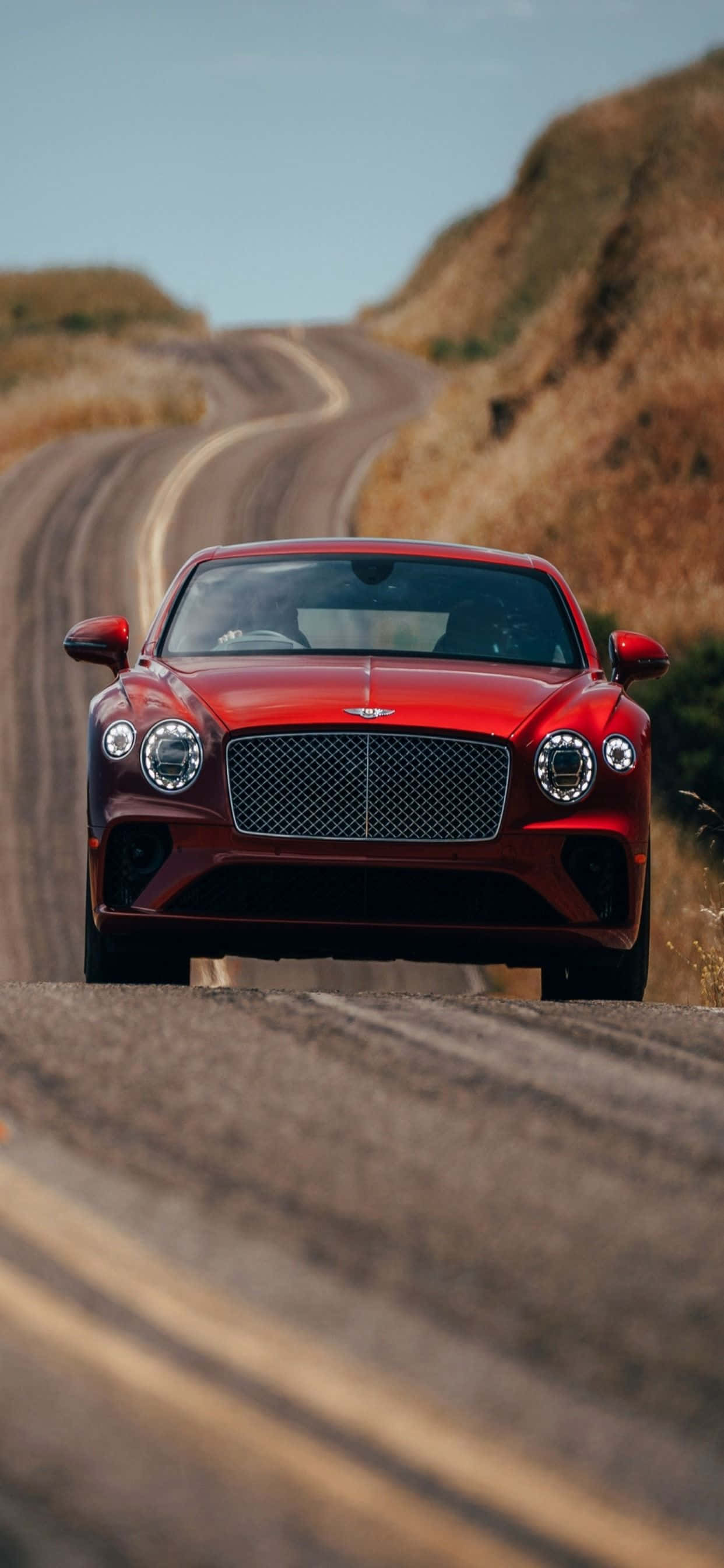 iPhone Xs Max Bentley 桌布 壁紙 — 一輛亮紅色的賓利 Bentayga 在晴朗藍天下沿著蜿蜒的沙漠道路駛向觀眾。