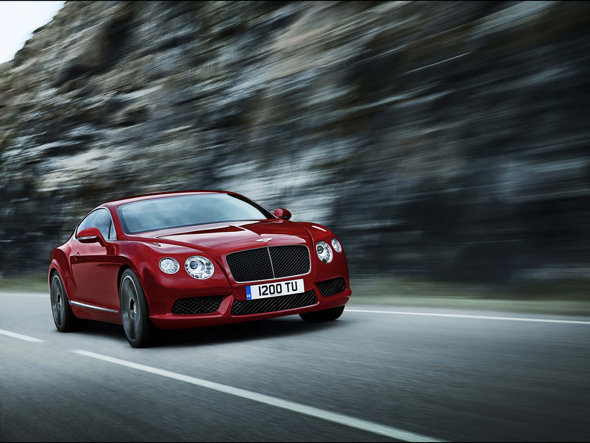 Bentley Continental Gt 桌布 壁紙 — 一輛亮紅色的賓利 Continental GT 在蜿蜒的山路上疾馳，模糊的岩石懸崖呼嘯而過。