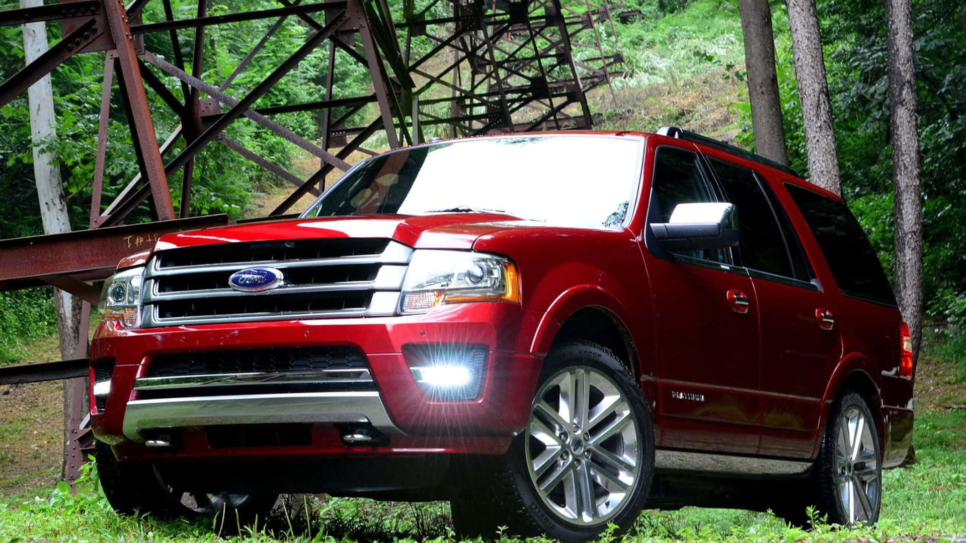 Ford Expedition 桌布 壁紙 — 一輛亮紅色福特 Expedition SUV 停放在生鏽金屬橋下，周圍是茂密的綠色森林。