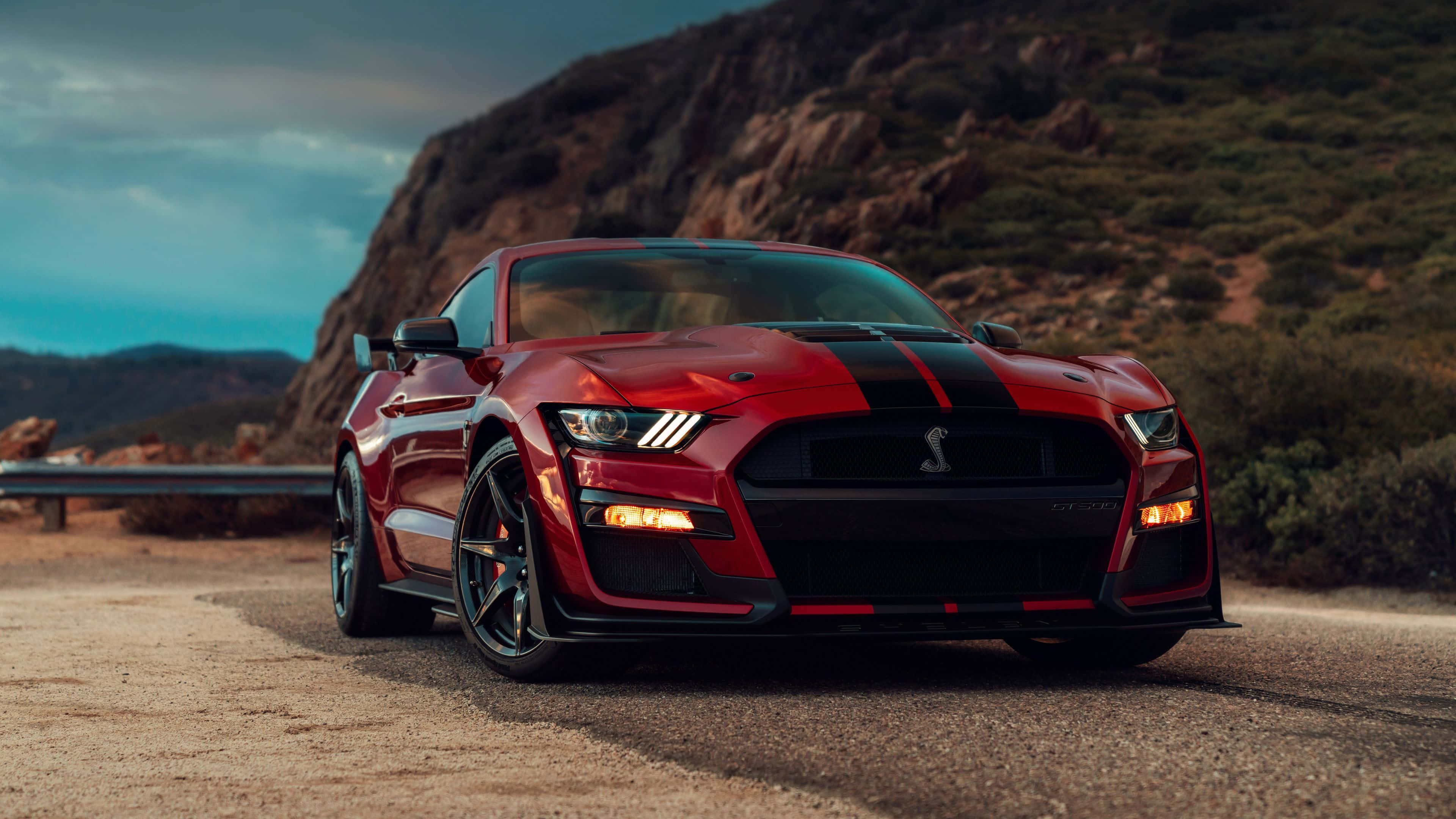 福特 桌布 壁紙 — 一輛亮紅色福特野馬Shelby GT500，帶有黑色賽車條紋，停泊在蜿蜒的山路上，天空陰沉。