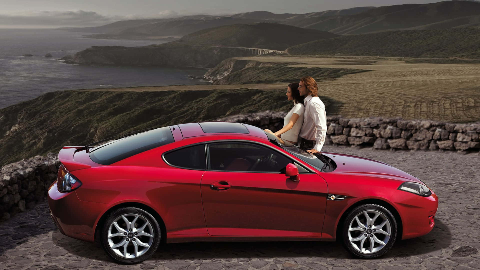 Hyundai Tiburon 桌布 壁紙 — 一輛亮紅色跑車停在石頭觀景點上，一對情侶靠在車頂，凝視著海岸景觀，有起伏的山丘和遠處的橋樑，天空柔和。