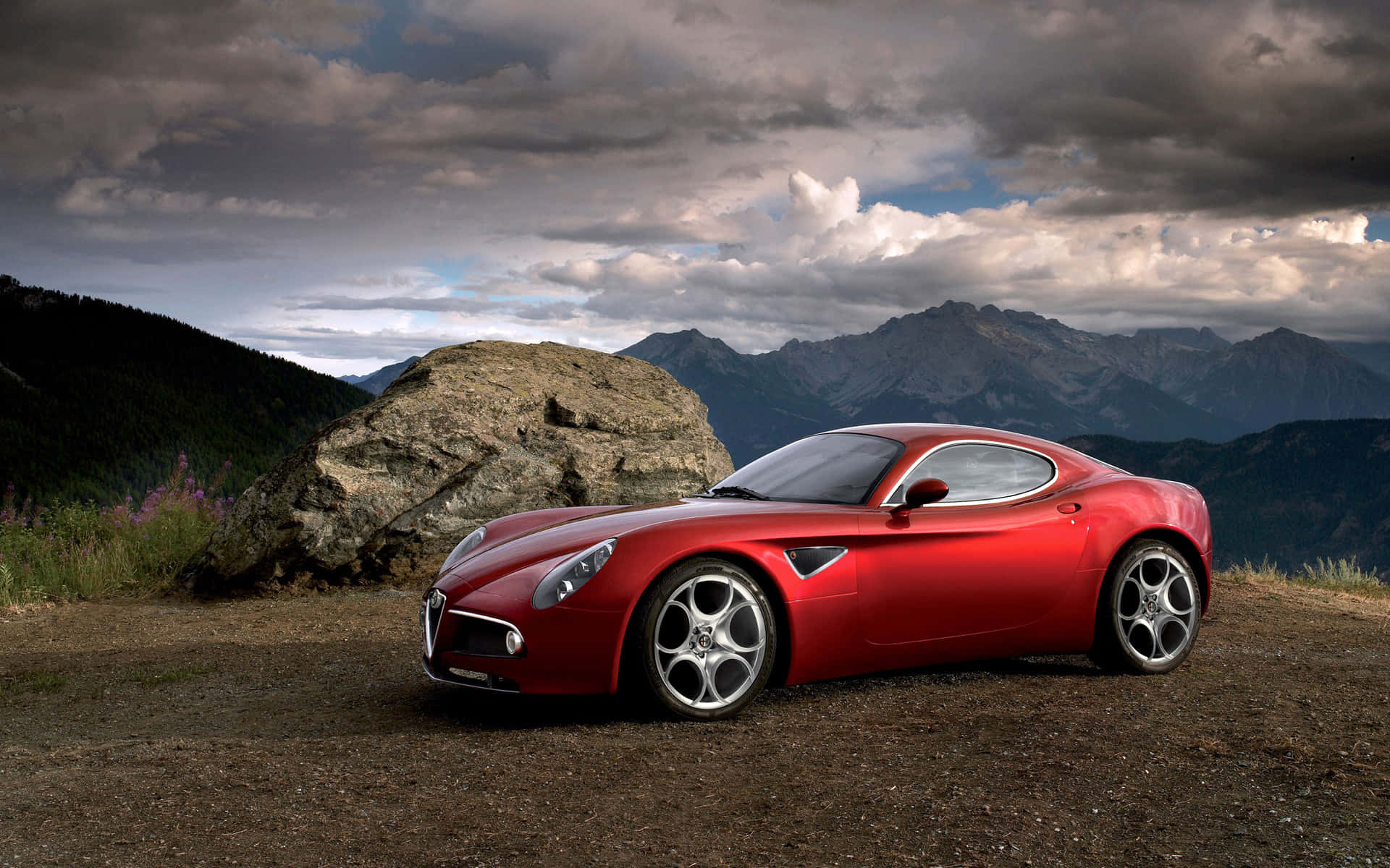 Alfa Romeo 8c Competizione 桌布 壁紙 — 一輛亮紅色跑車停泊在山脊上，背景是戲劇性的多雲天空和崎嶇的山峰。