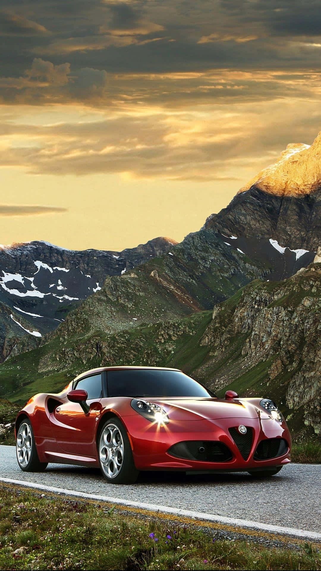 Alfa Romeo 4c 桌布 壁紙 — 一輛亮紅色跑車在日落時分行駛在山路上，背景是白雪覆蓋的山峰和金色的雲彩。