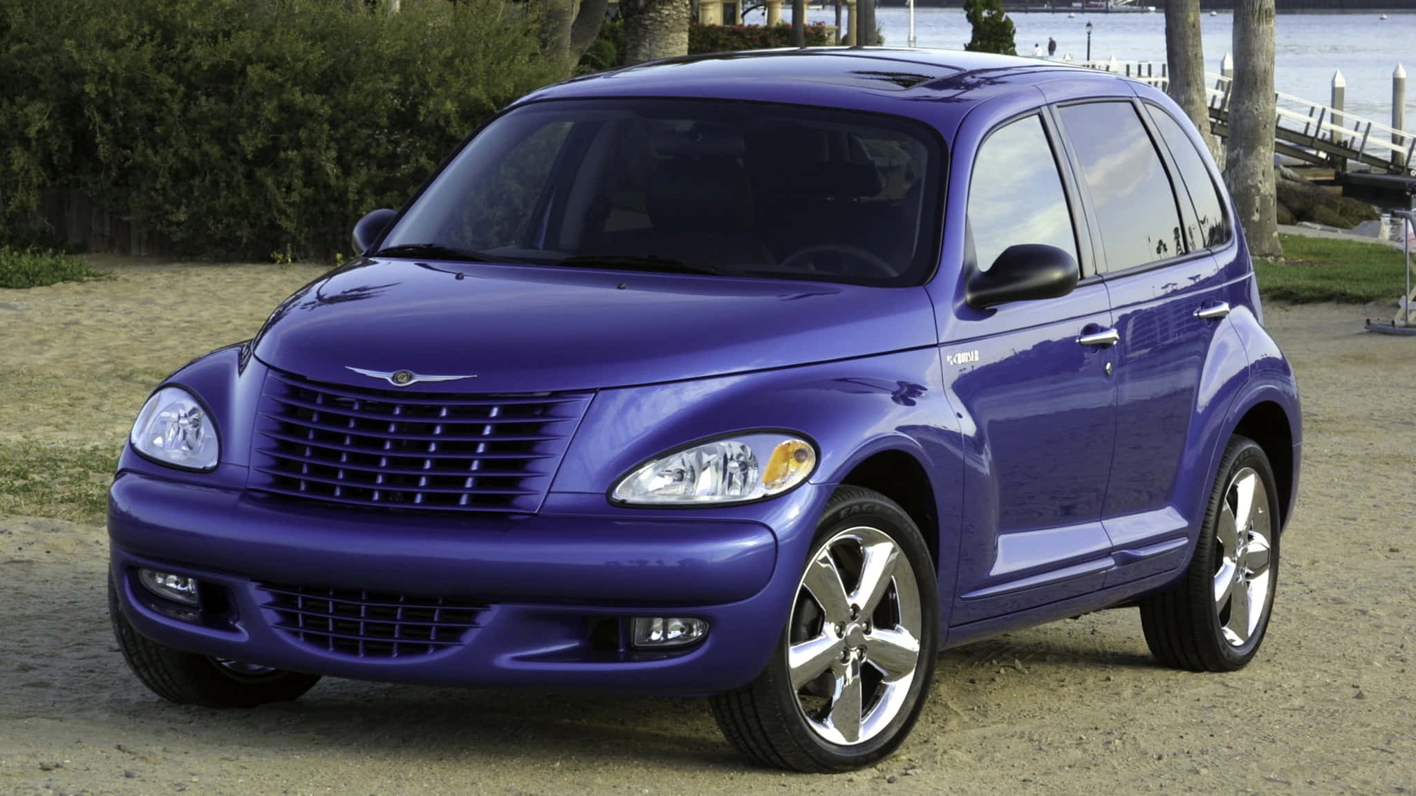 Chrysler Pt Cruiser 桌布 壁紙 — 一輛亮紫色的PT Cruiser停放在近水的碎石上，背景有樹木和碼頭。