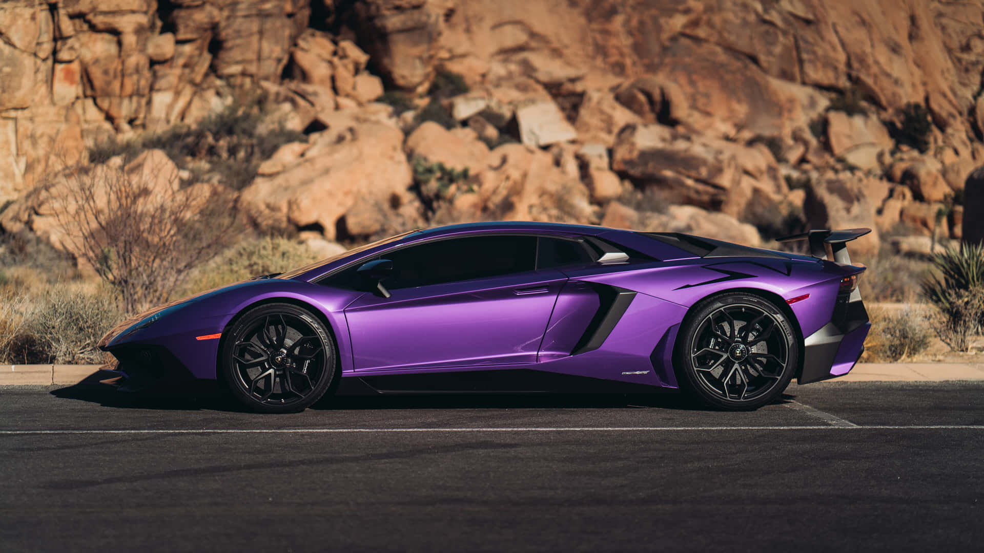 Lamborghini purple 壁紙 — 一輛亮紫色藍寶堅尼 Aventador SVJ 停在柏油路上，背景是崎嶇的棕色岩層。