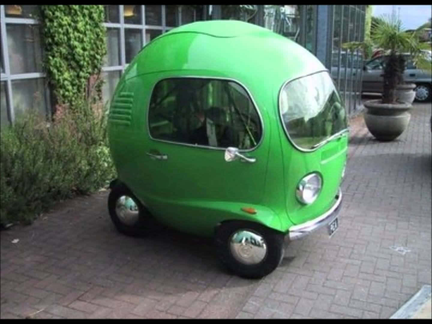 cars 桌布 壁紙 — 一輛亮綠色蛋形復古車停在磚砌人行道上，旁邊是玻璃建築和盆栽植物。