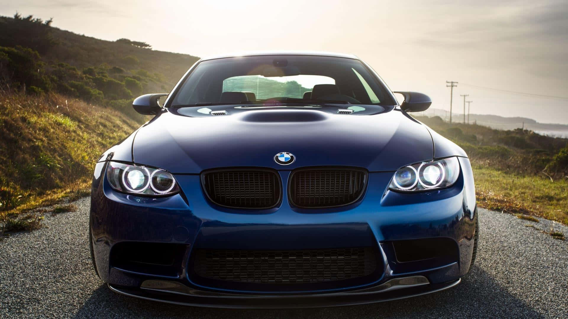 1920x1080 Bmw 桌布 壁紙 — 一輛亮藍色BMW M3跑車在沿海公路上向前行駛，金色陽光照射在引擎蓋和車頭燈上。