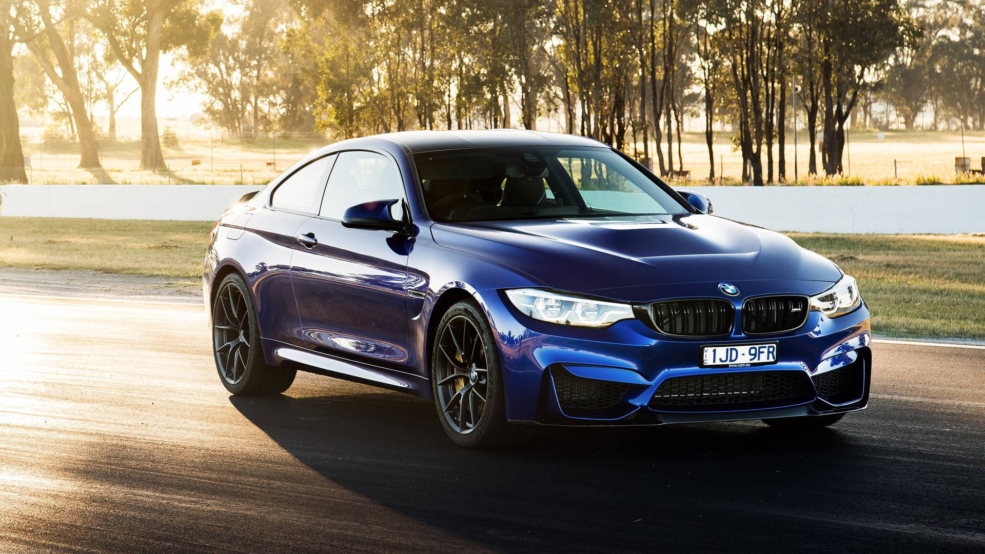 日落時分賽道上的藍色BMW M4雙門跑車，背景有樹木和護欄。