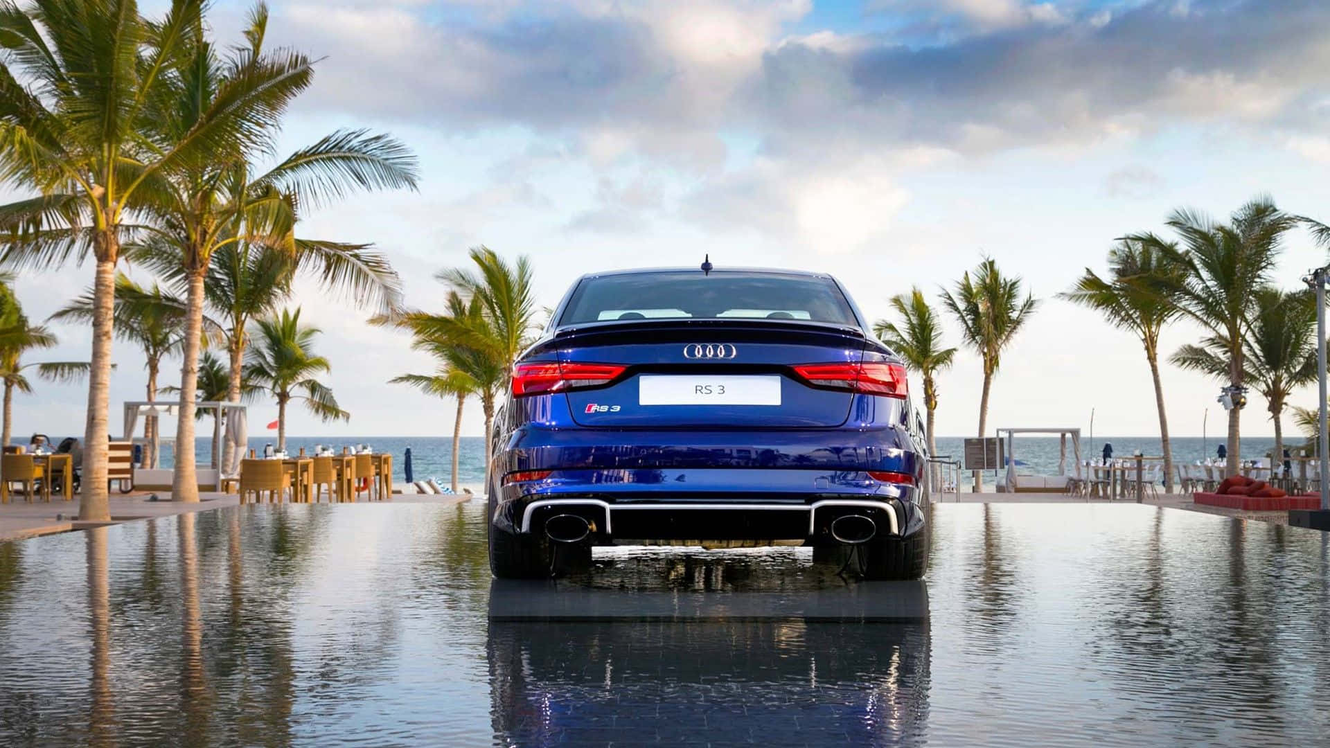 Audi Rs3 桌布 壁紙 — 一輛亮藍色奧迪RS3房車停放在一個反射的無邊際泳池中央，背景有棕櫚樹和海洋，天空部分多雲。