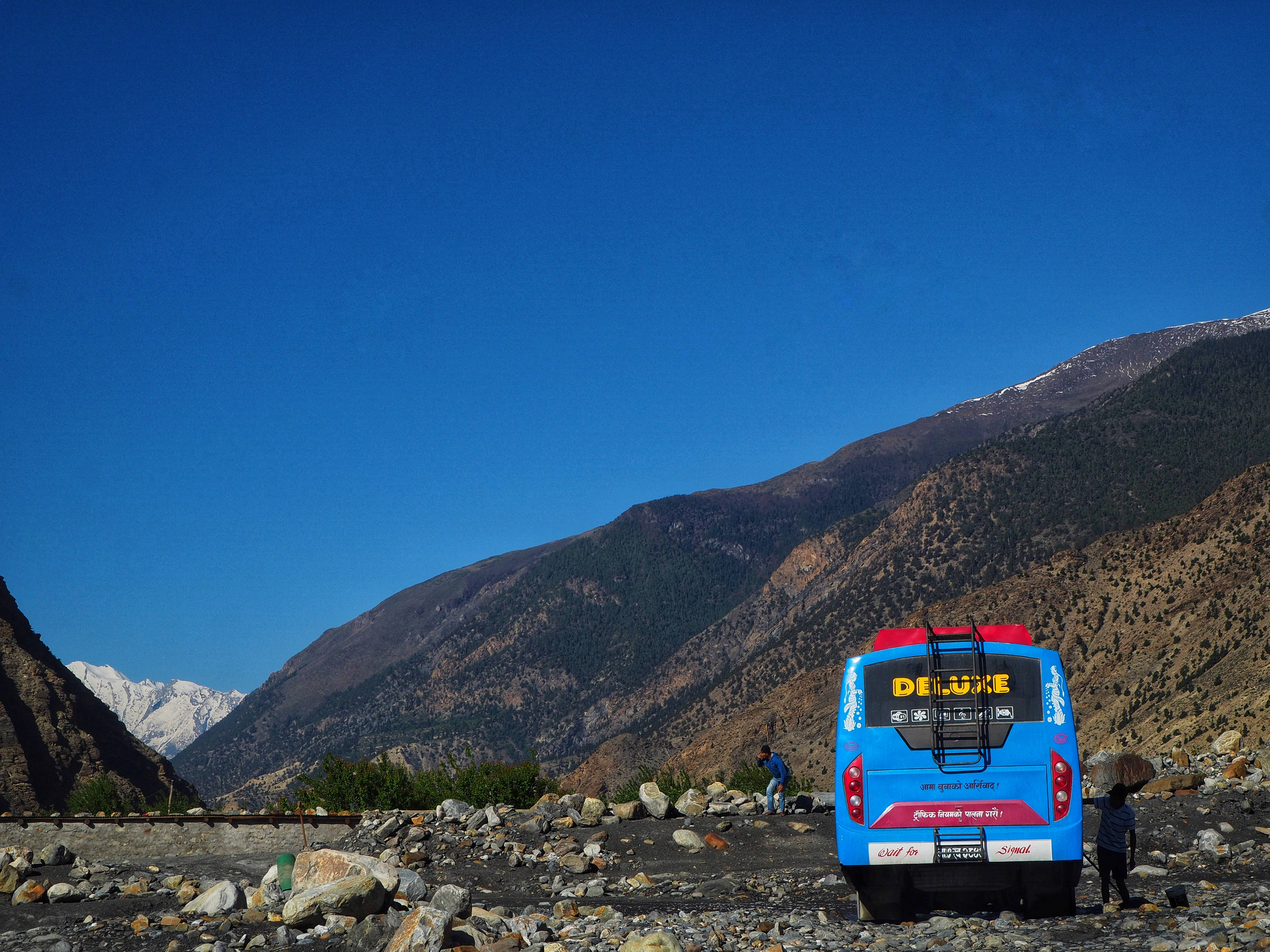 Nepal 桌布 壁紙 — 一輛亮藍色巴士停泊在岩石山路上，背景是白雪覆蓋的山峰和清澈的藍天。