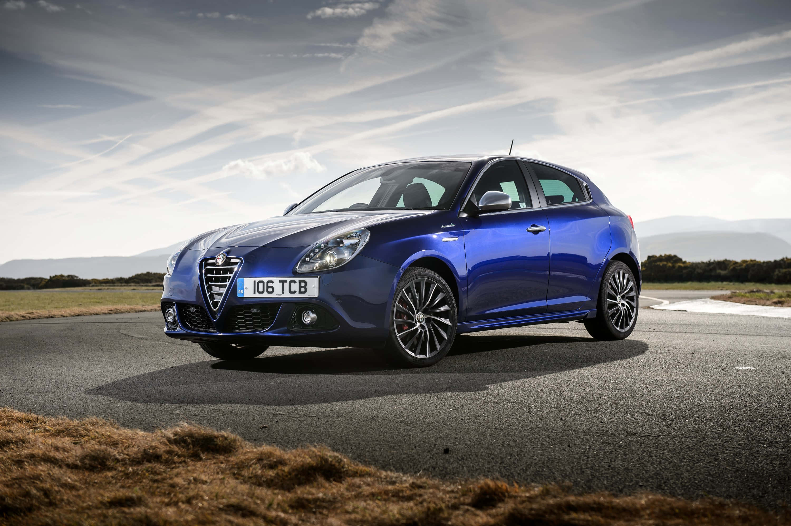 Alfa Romeo Giulietta 桌布 壁紙 — 一輛亮藍色愛快羅密歐Giulietta停泊在蜿蜒道路上，遠方山丘在陰天下。