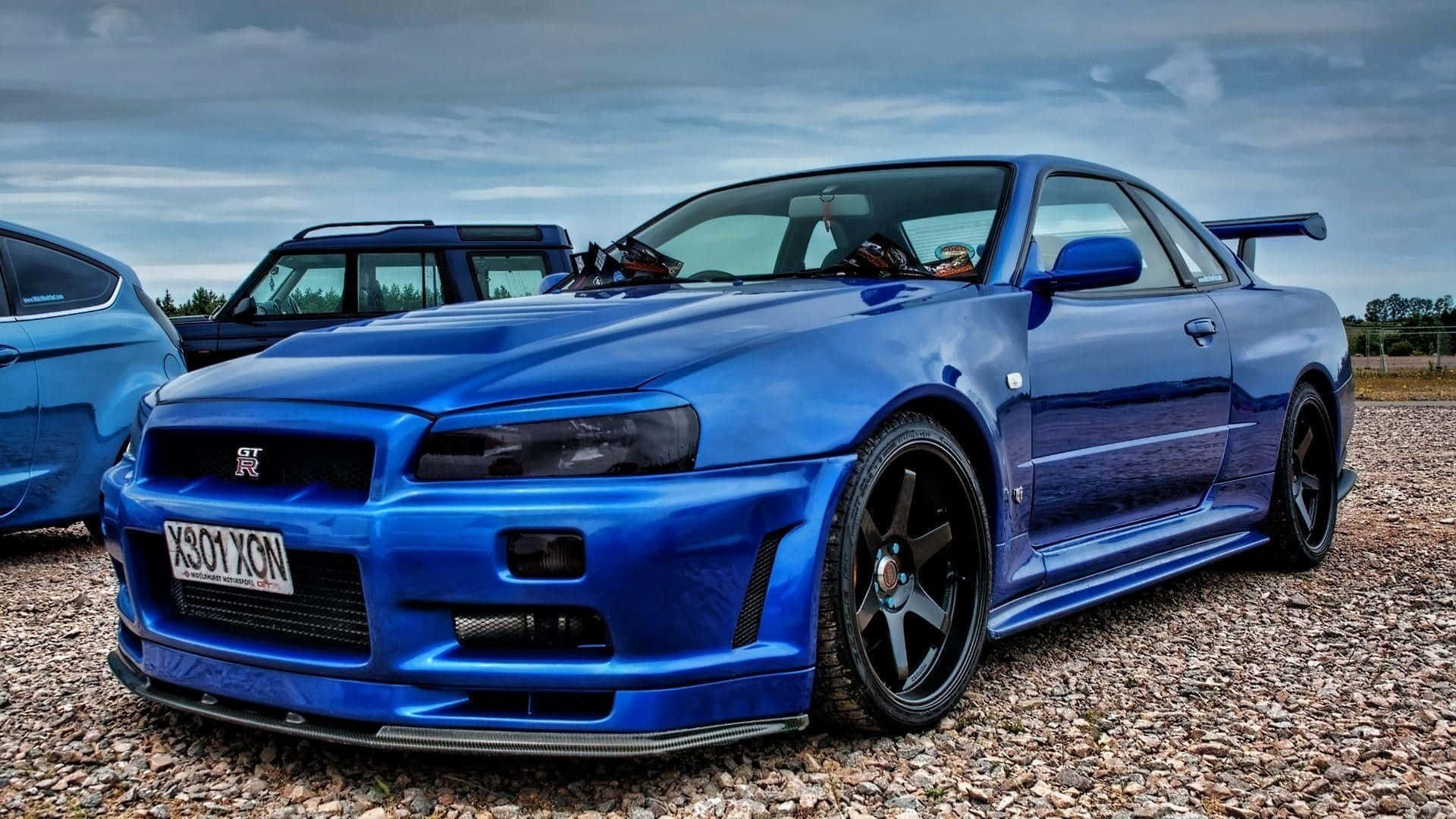  壁紙 — 一輛亮藍色日產Skyline GT-R跑車停放在碎石地上，天空多雲，附近有另一輛藍色汽車。
