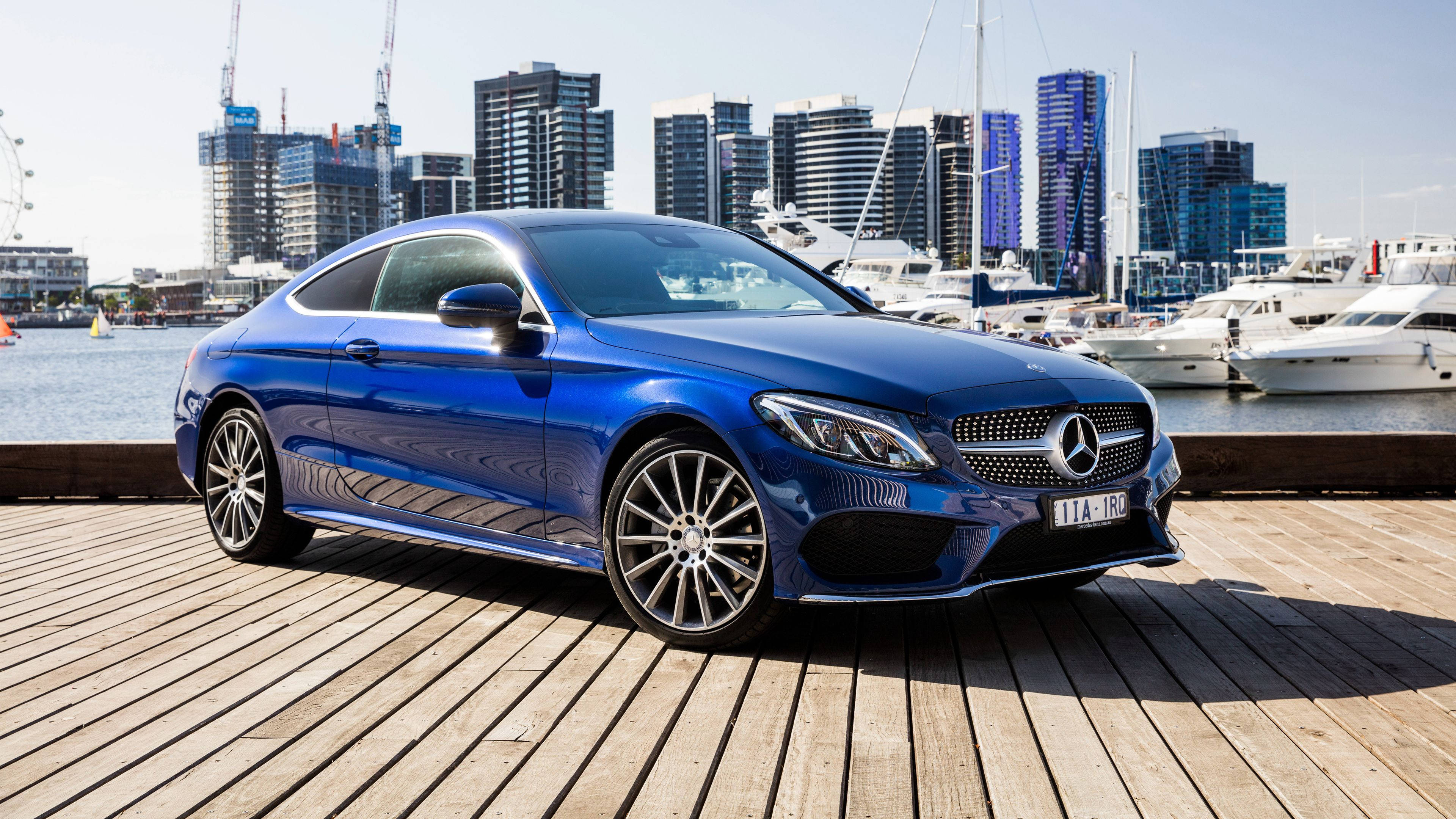 Mercedes Benz C300 桌布 壁紙 — 一輛亮藍色梅賽德斯轎跑車停泊在木製碼頭上，背景有遊艇和現代城市天際線。