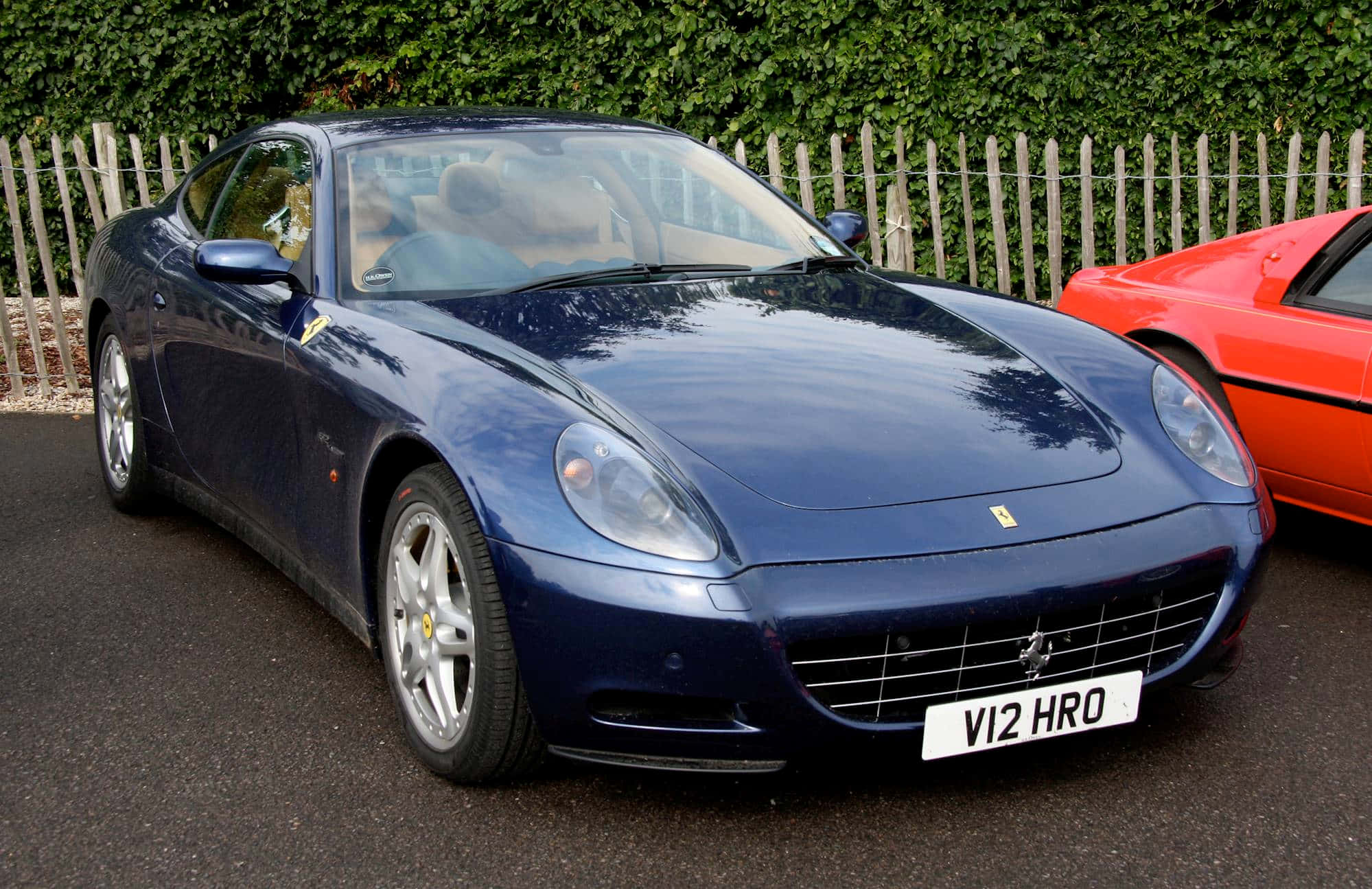 Ferrari 612 Scaglietti 桌布 壁紙 — 一輛亮藍色法拉利跑車停泊在柏油路上，旁邊有一輛紅色汽車，後面有綠色樹籬。