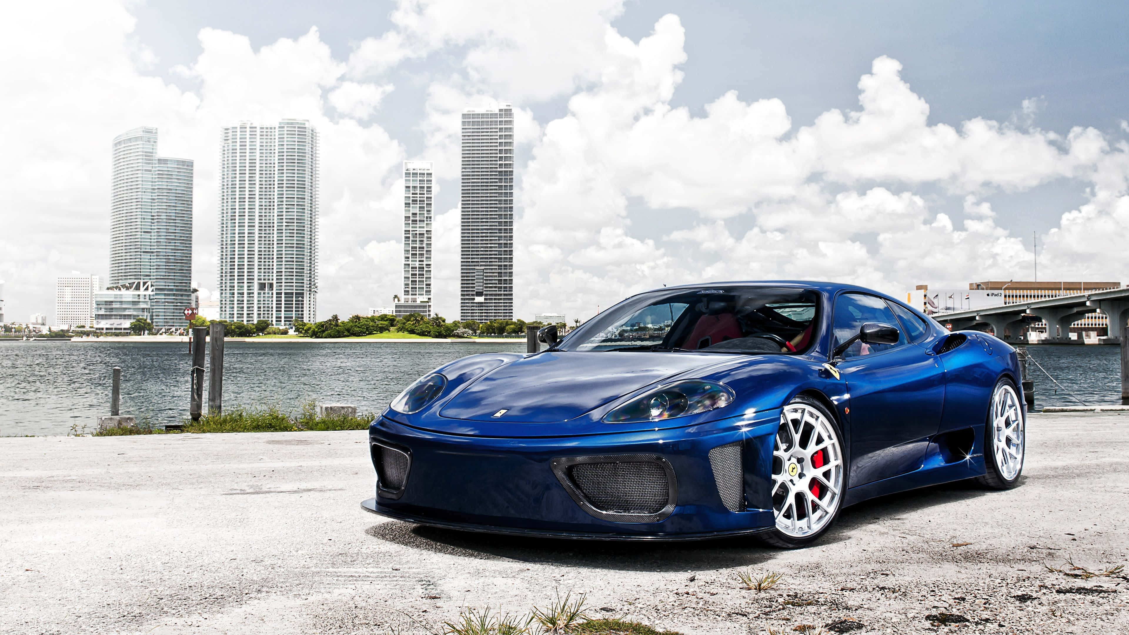 Ferrari 360 Modena 桌布 壁紙 — 一輛亮藍色法拉利跑車停泊在海濱碼頭，背景是現代摩天大樓和橋樑，天空部分多雲。