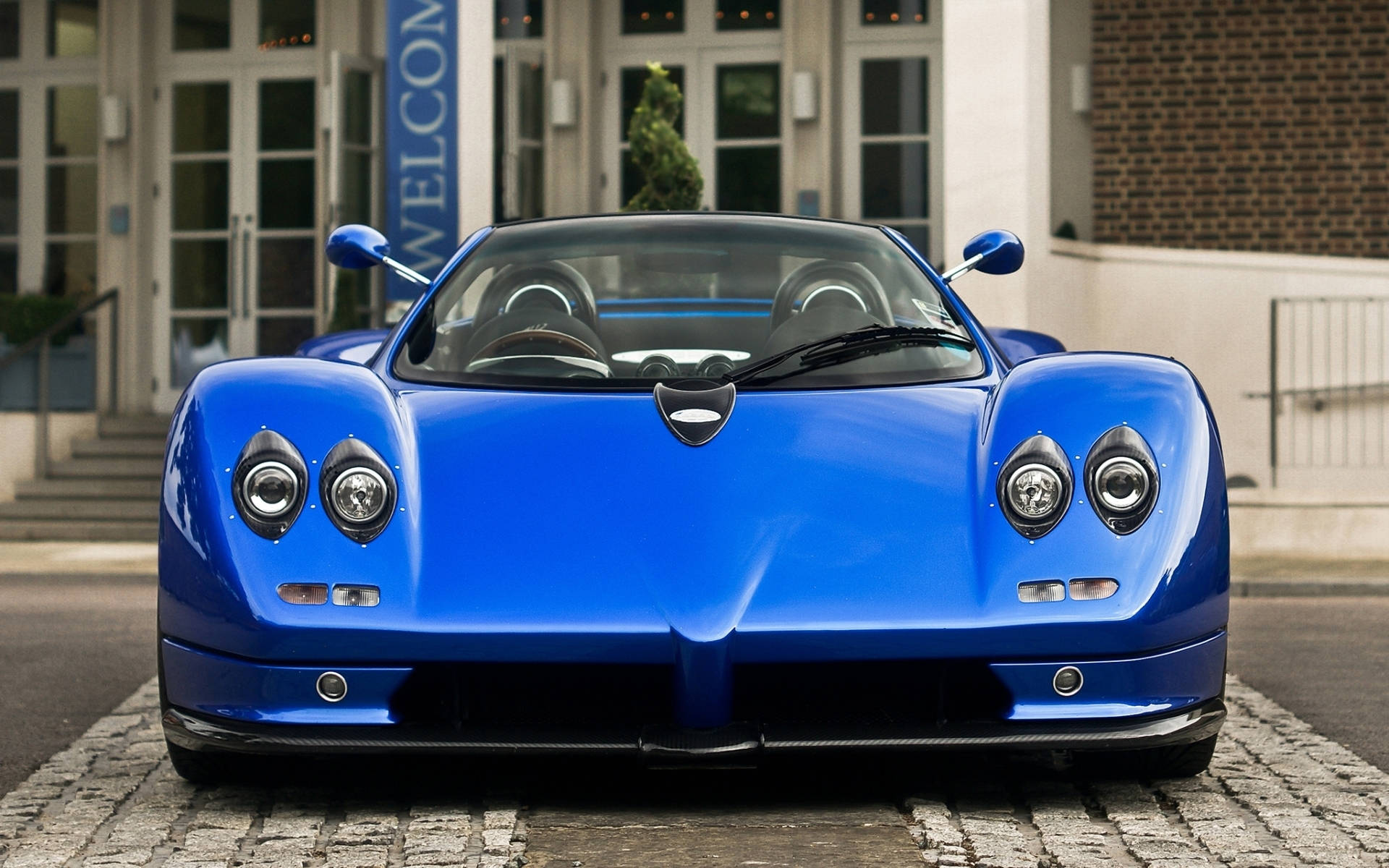pagani iPhone 桌布 壁紙 — 一輛亮藍色的Pagani Zonda跑車停在現代建築前的鵝卵石上。