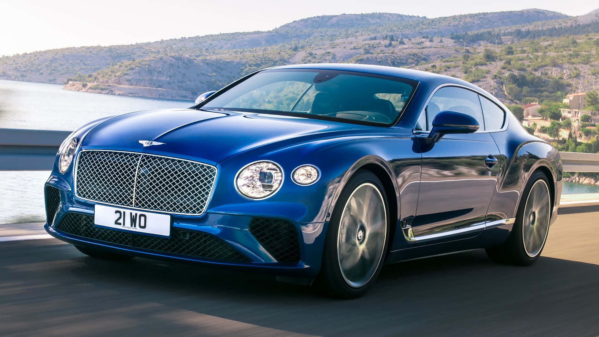 Best Bentley 桌布 壁紙 — 一輛亮藍色賓利 Continental GT 沿著海岸道路行駛，背景有山丘和水面。