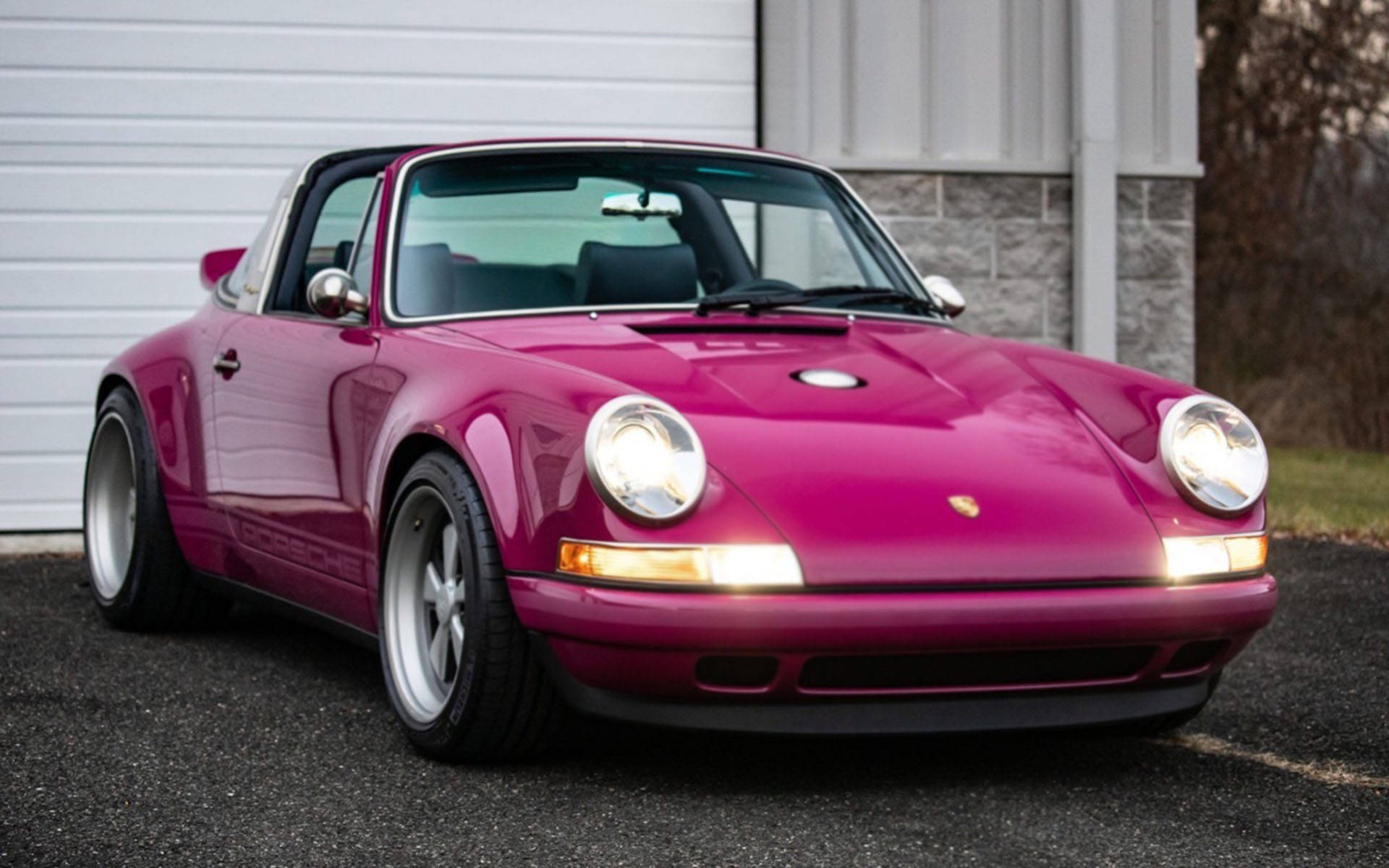 Singer 桌布 壁紙 — 一輛亮面洋紅色保時捷911 Targa停在白色車庫門前的瀝青路上，車頭燈亮著。