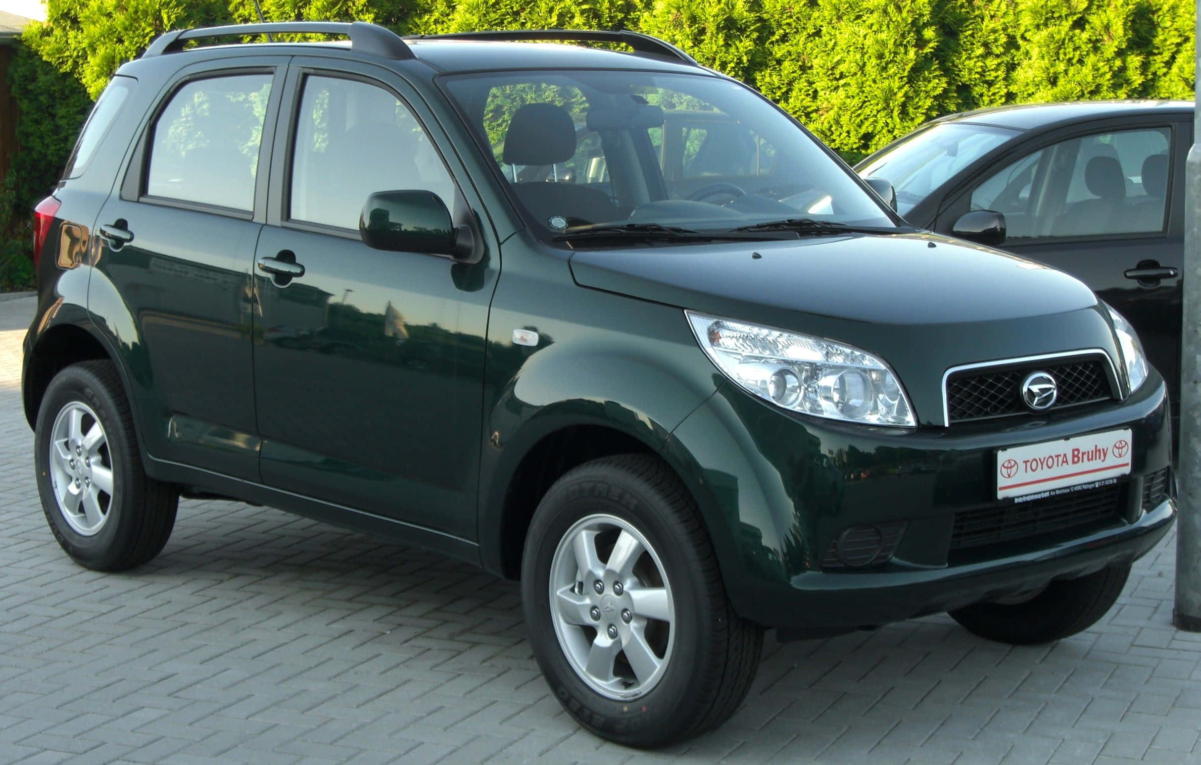 Daihatsu 桌布 壁紙 — 一輛亮面深綠色緊湊型SUV停在鋪砌的停車場上，背景有茂密的綠色灌木叢。