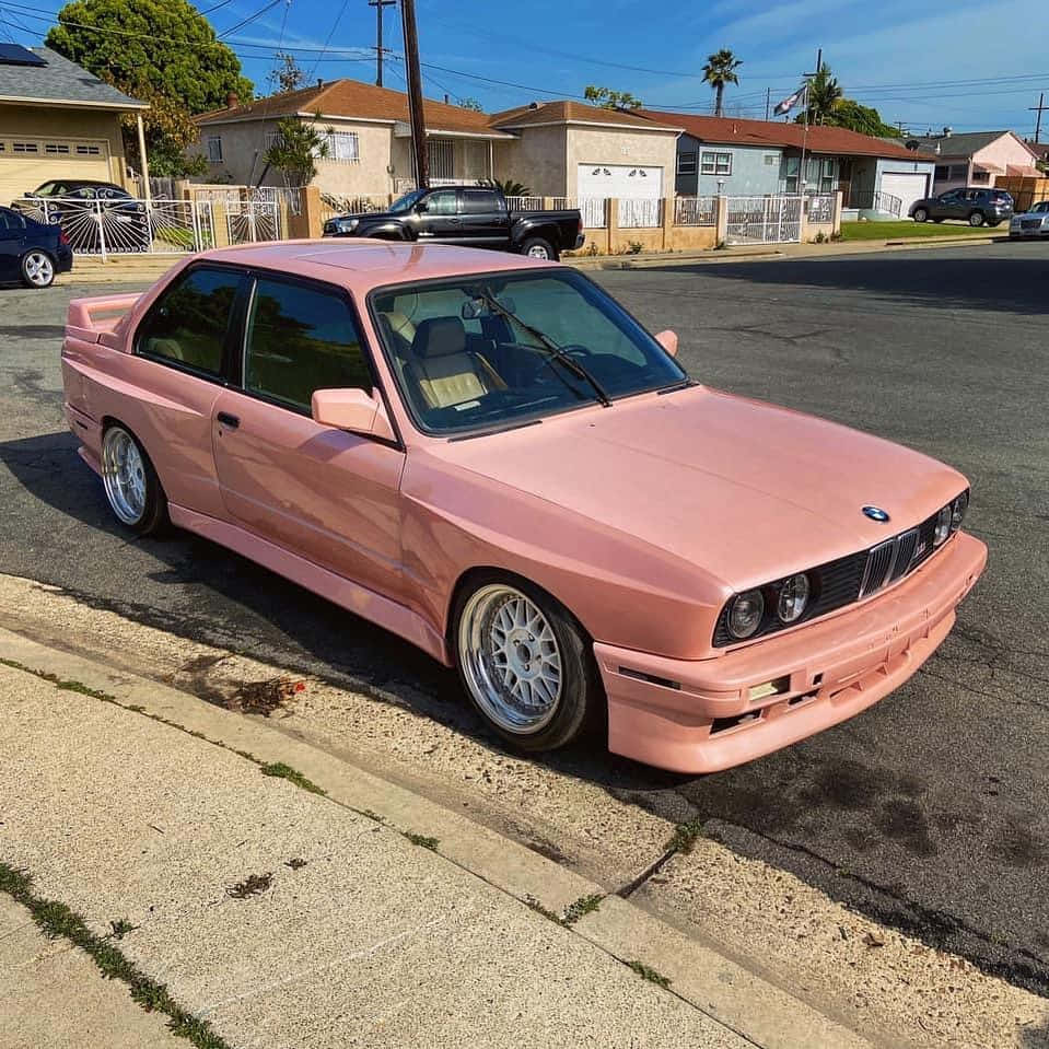 BMW 動圖 桌布 壁紙 — 一輛亮面粉紅色BMW E30雙門跑車停放在陽光明媚的郊區街道上，背景有棕櫚樹和房屋。