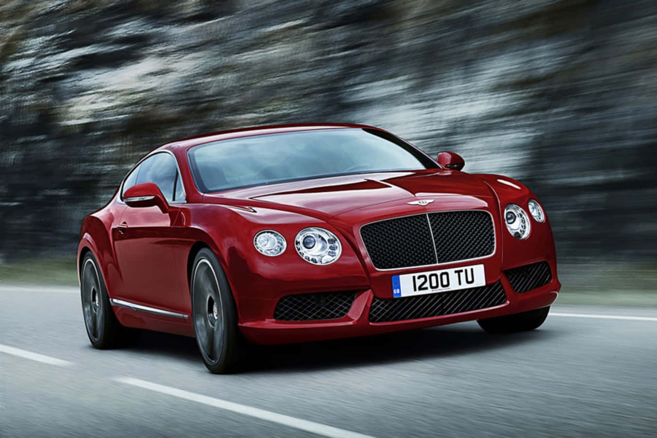 賓利 桌布 壁紙 — 一輛亮面紅色 Bentley Continental GT 在蜿蜒的道路上疾馳，背景是模糊的岩石懸崖。