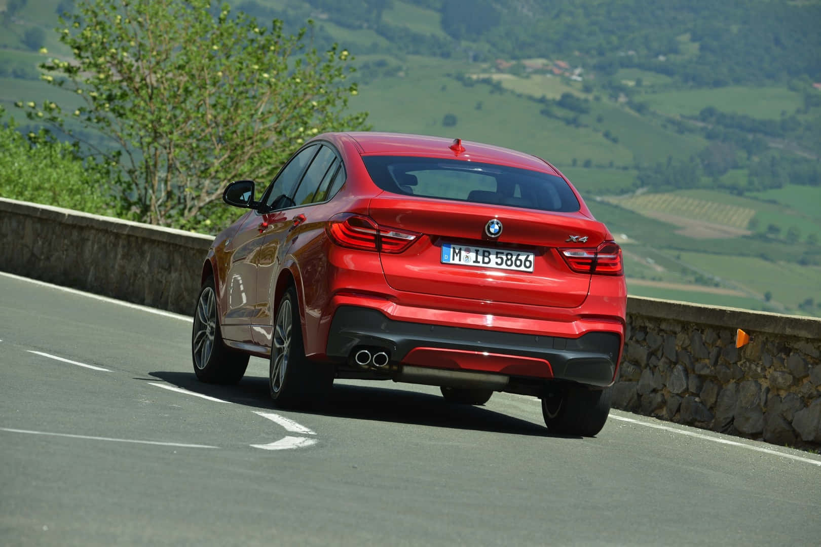 Bmw X4 桌布 壁紙 — 一輛亮面紅色BMW X4在明媚日光下行駛於蜿蜒的山路上，兩旁是翠綠的山丘和石牆。