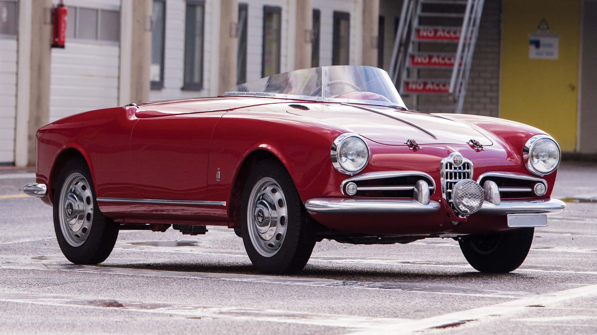 Alfa Romeo Spider 桌布 壁紙 — 一輛亮面紅色復古愛快羅密歐Spider敞篷車停放在濕瀝青地上，背景有工業建築。