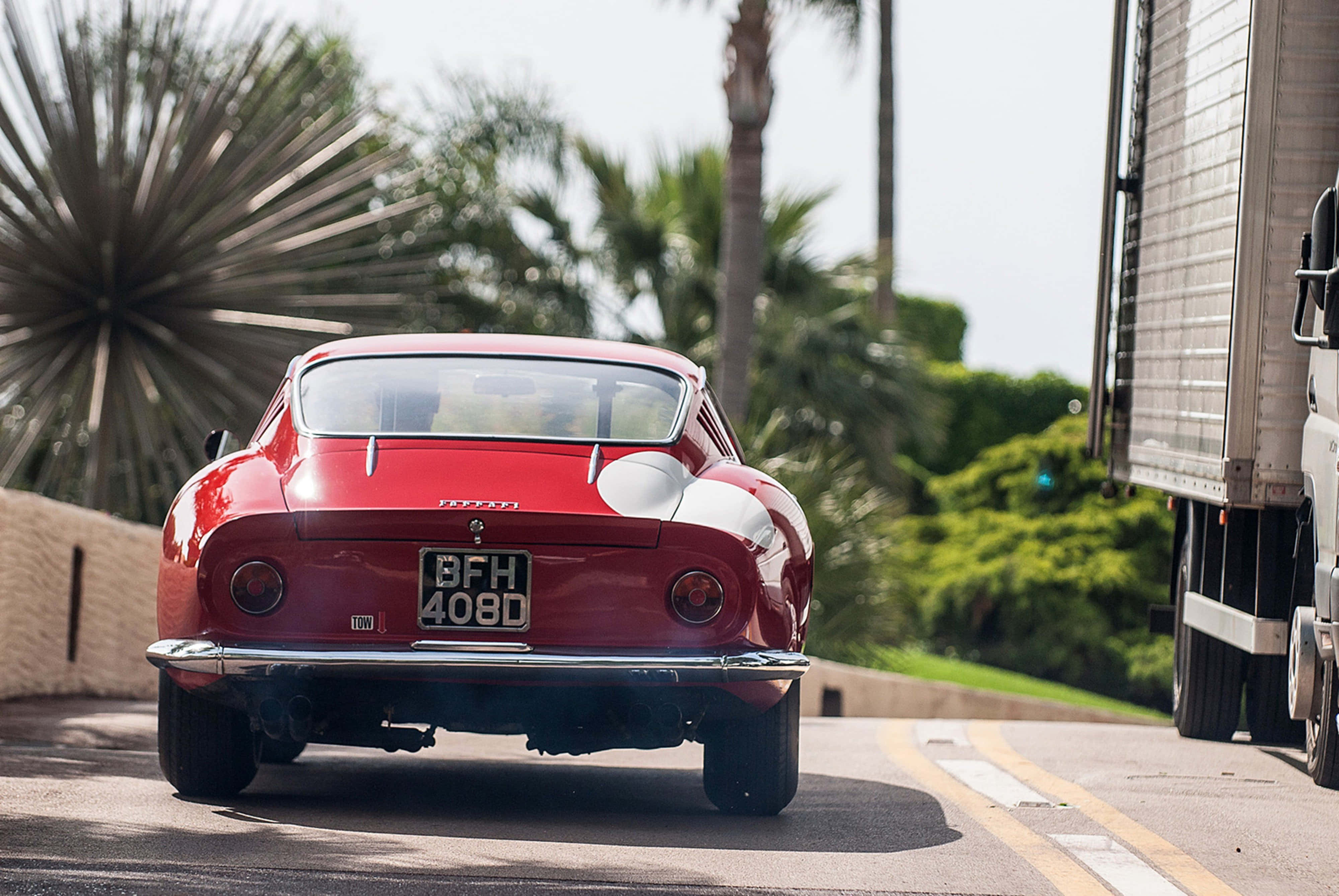 Vintage Ferrari 桌布 壁紙 — 一輛亮面紅色復古法拉利250 GT在陽光明媚的道路上行駛，兩旁有棕櫚樹和一輛大型卡車。