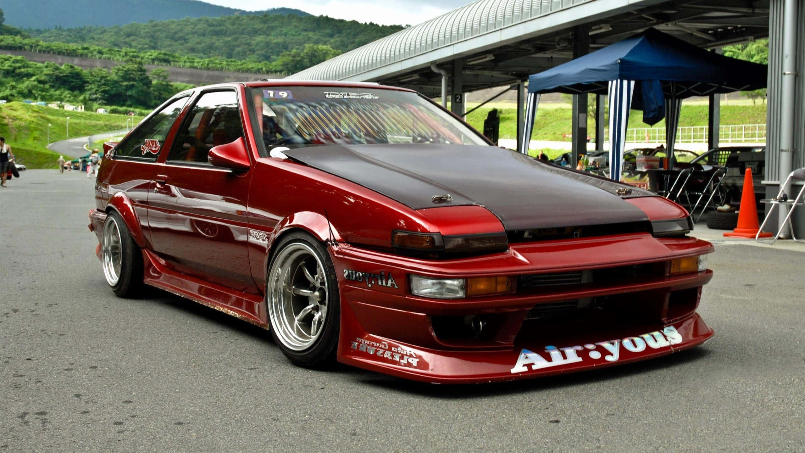 JDM 桌布 壁紙 — 一輛亮面紅色改裝AE86跑車，配備寬體車身套件和訂製輪圈，停在賽車場維修區。