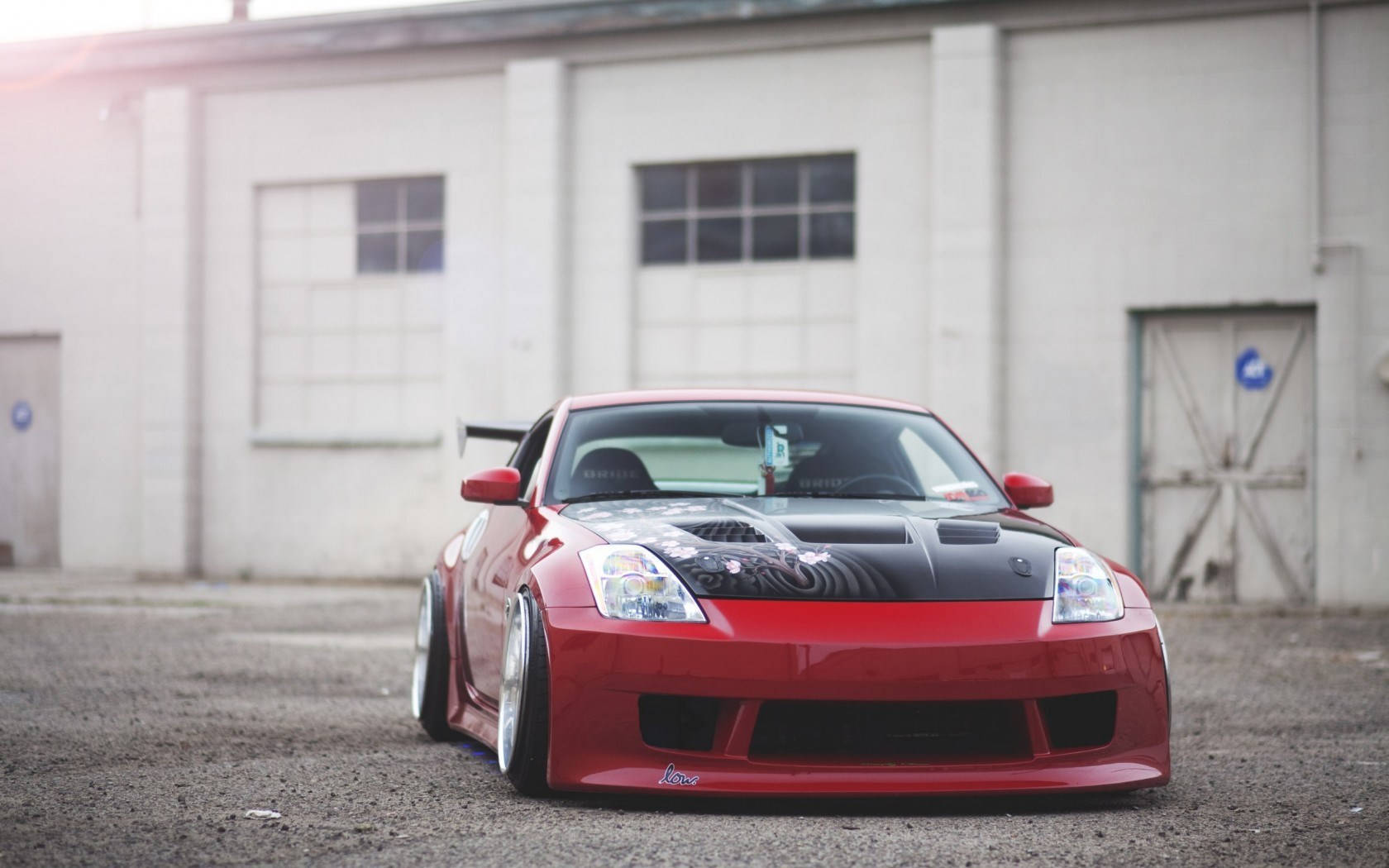 Nissan 350z 桌布 壁紙 — 一輛亮面紅色改裝跑車，黑色引擎蓋和客製化輪圈，停放在風化的工業建築前。