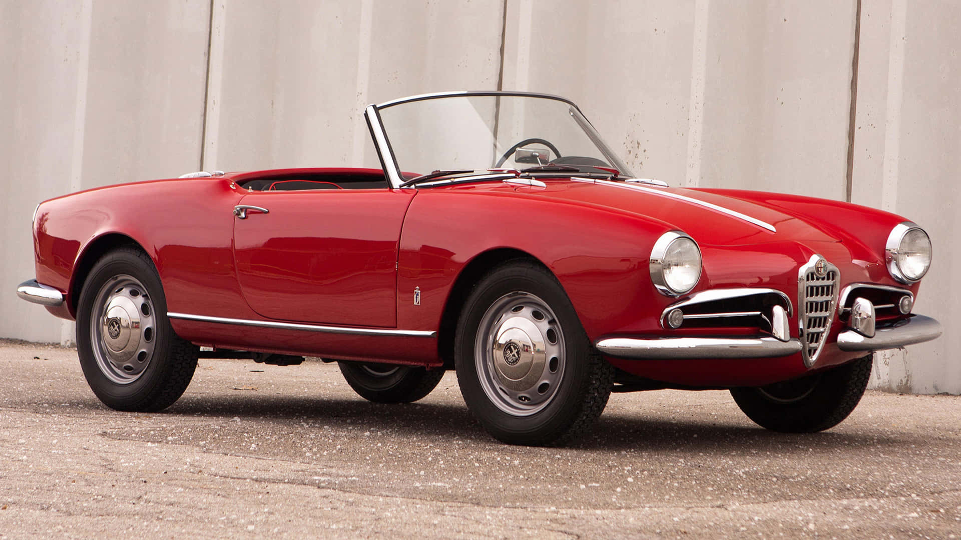 Alfa Romeo Spider 桌布 壁紙 — 一輛亮面紅色經典愛快羅密歐Spider敞篷車停泊在碎石上，背景是波浪形金屬牆。