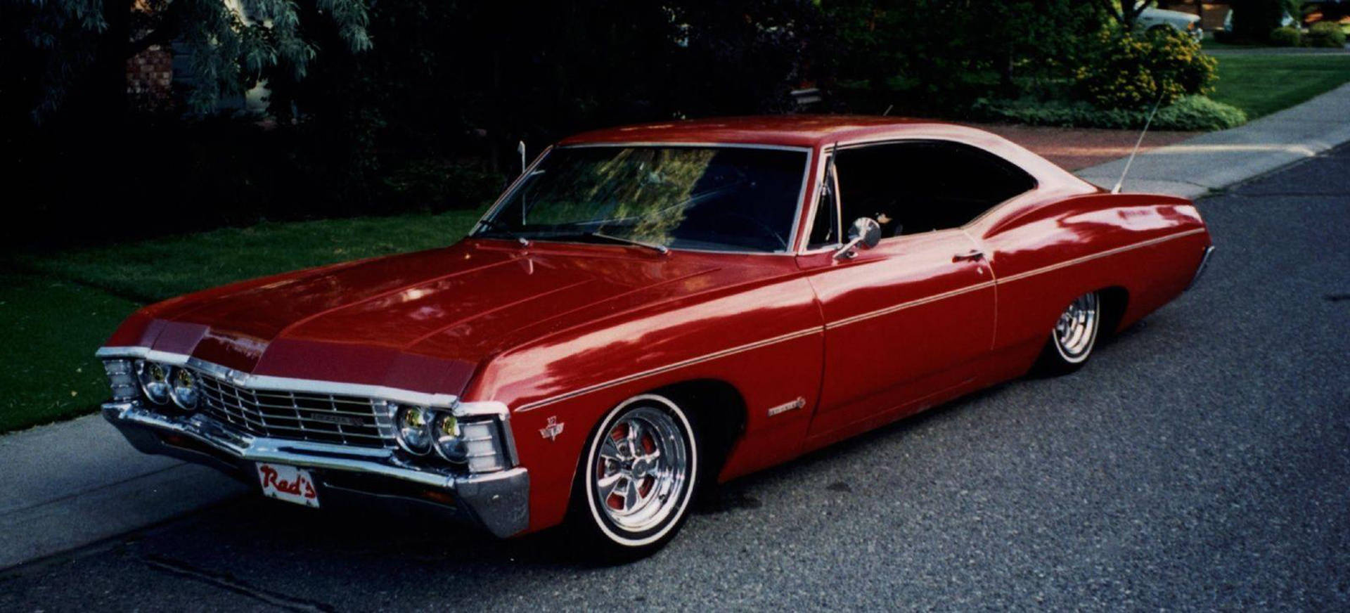 chevrolet impala 1967 桌布 壁紙 — 一輛亮面紅色經典肌肉車，帶有鍍鉻細節，停泊在黃昏的郊區街道上。