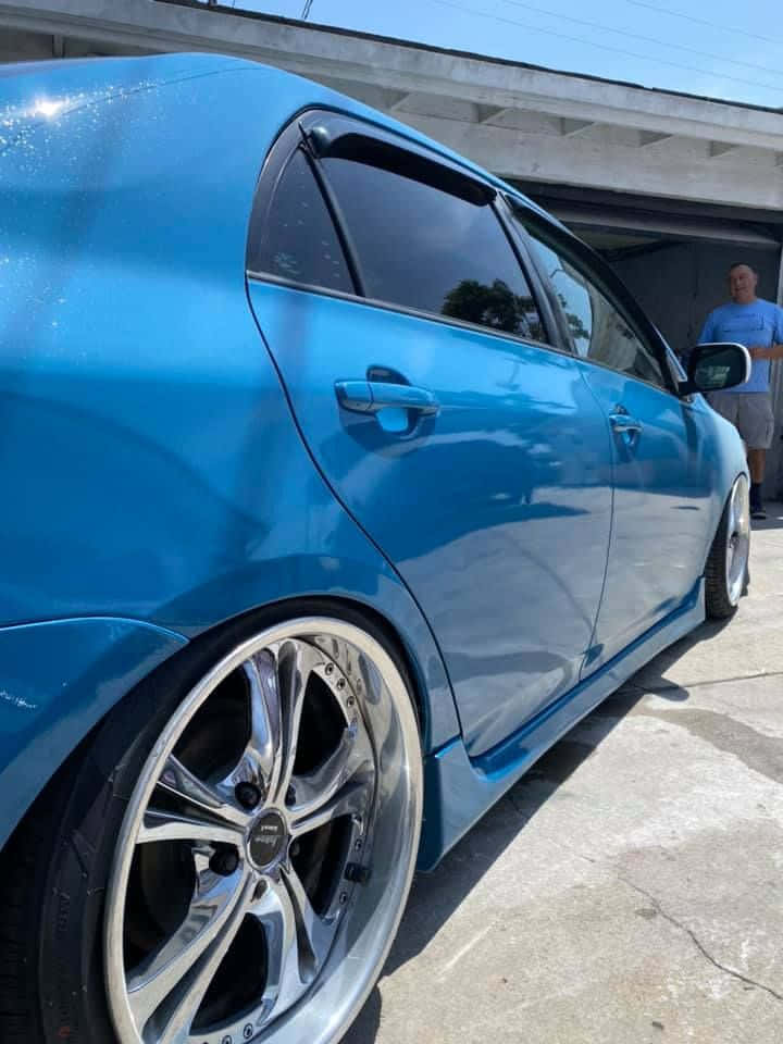 Azure Blue 桌布 壁紙 — 一輛亮面藍色改裝轎車配備定制鍍鉻輪圈，停泊在混凝土高架橋下，附近站著一名男子。