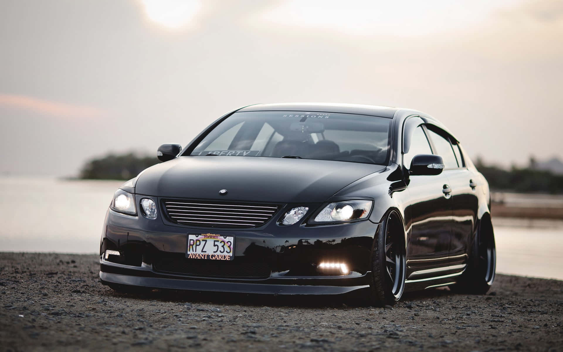 Lexus Gs 桌布 壁紙 — 一輛亮面黑色改裝轎車，帶有發光車頭燈，停在日落時的礫石岸邊。