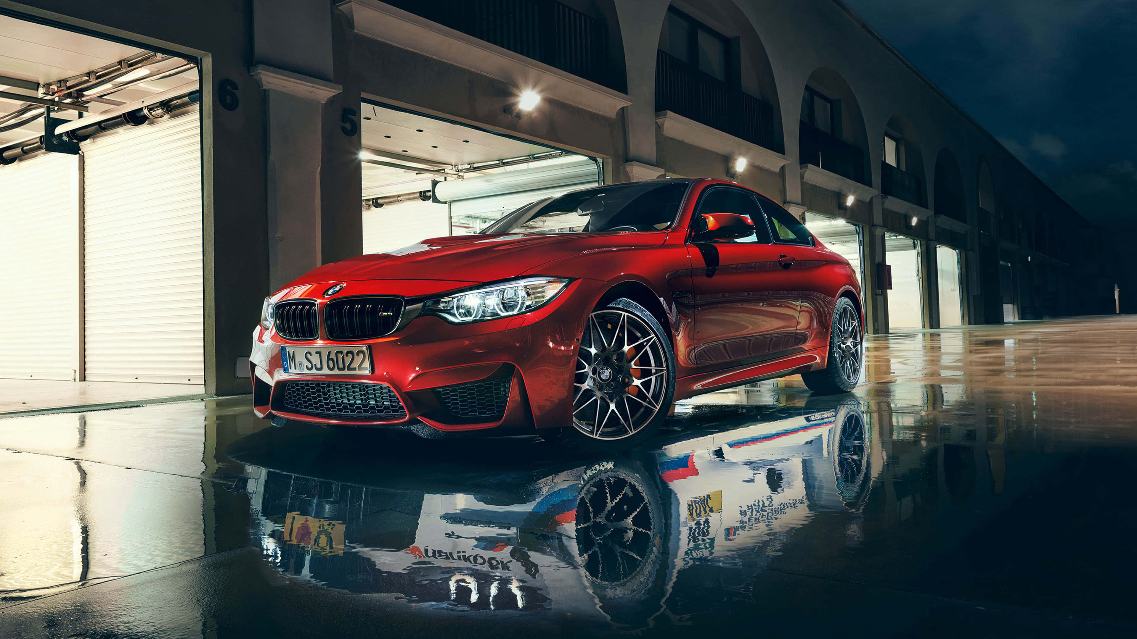BMW 動圖 桌布 壁紙 — 一輛亮麗的紅色BMW M4轎跑車在夜燈下閃耀，其倒影在現代車庫外的濕漉漉路面上閃爍。