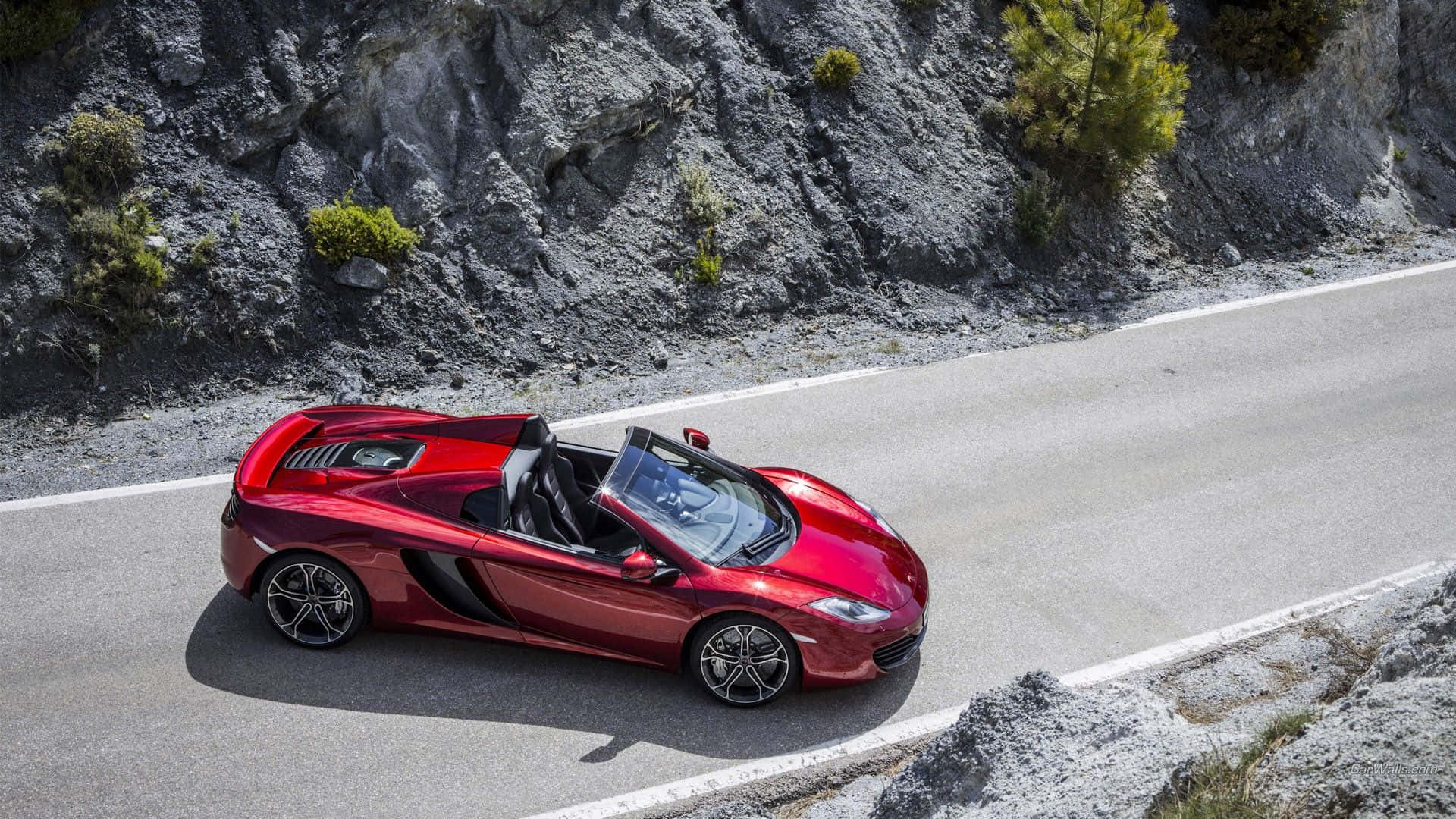 Mclaren F1 桌布 壁紙 — 一輛亮麗的紅色McLaren跑車在明亮的日光下行駛在蜿蜒的山路上，兩旁是岩石峭壁和稀疏的綠植。
