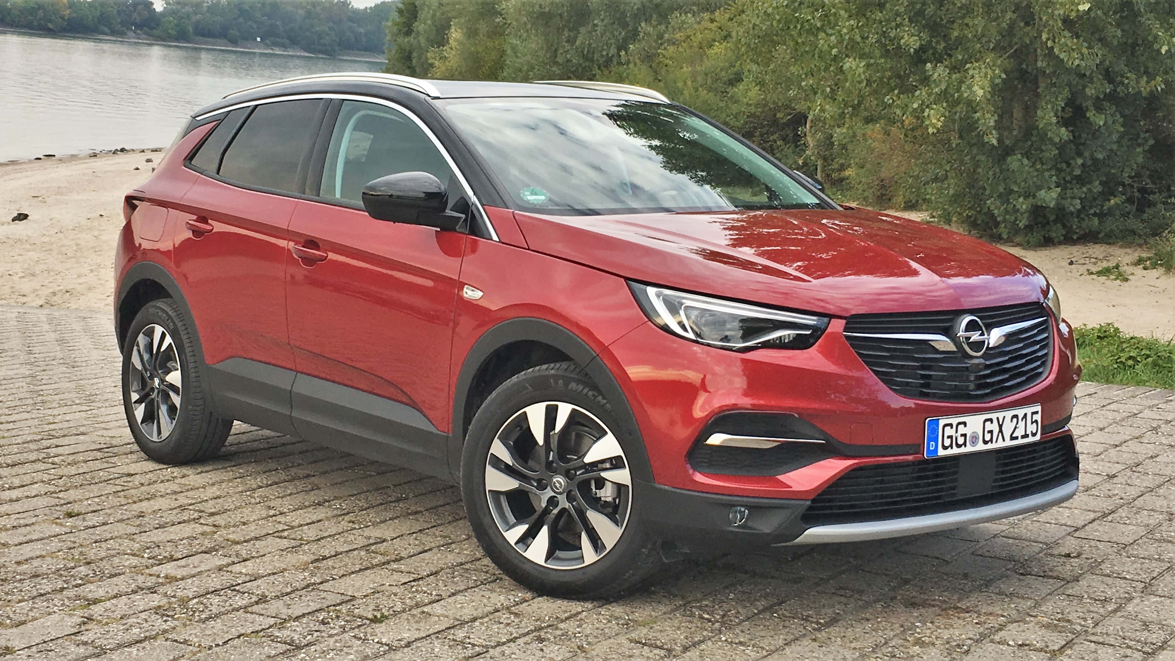 Opel Grandland X 桌布 壁紙 — 一輛亮麗的紅色Opel Grandland X SUV停泊在平靜河邊的鵝卵石小徑上，背景有綠樹。