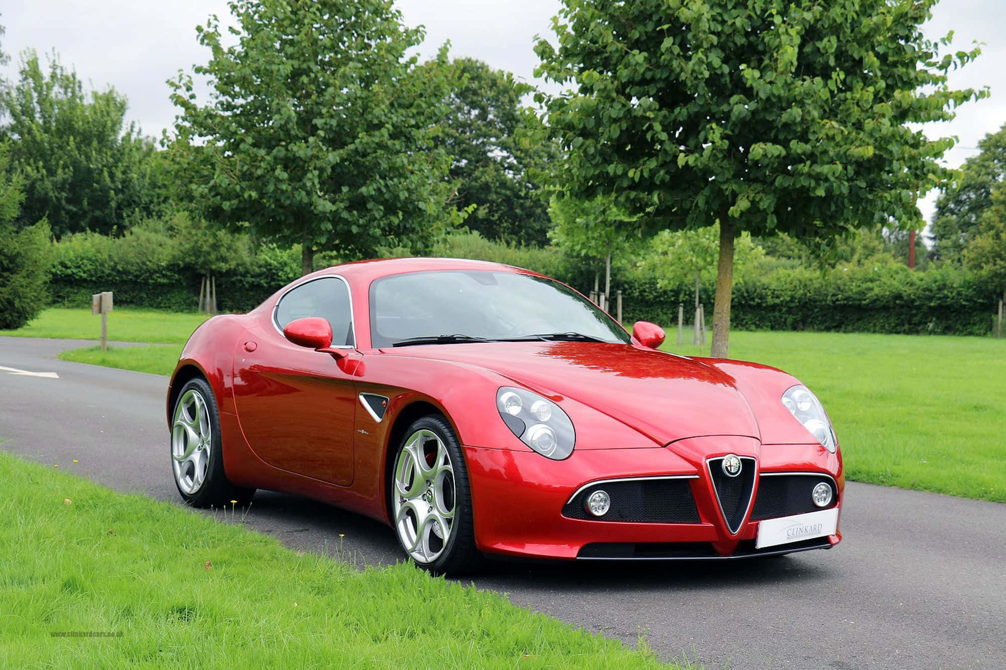 Alfa Romeo 8c Competizione 桌布 壁紙 — 一輛亮麗的紅色愛快羅密歐跑車停泊在蜿蜒的道路旁，旁邊是茂密的綠草和枝葉繁茂的樹木，天空多雲。