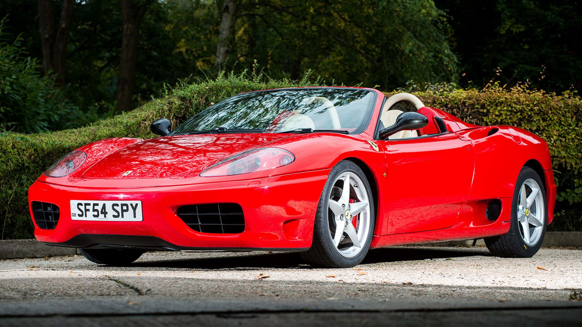 Ferrari 360 Modena 桌布 壁紙 — 一輛亮麗的紅色法拉利360 Spider停泊在碎石上，背景是茂密的綠色樹籬和樹木。