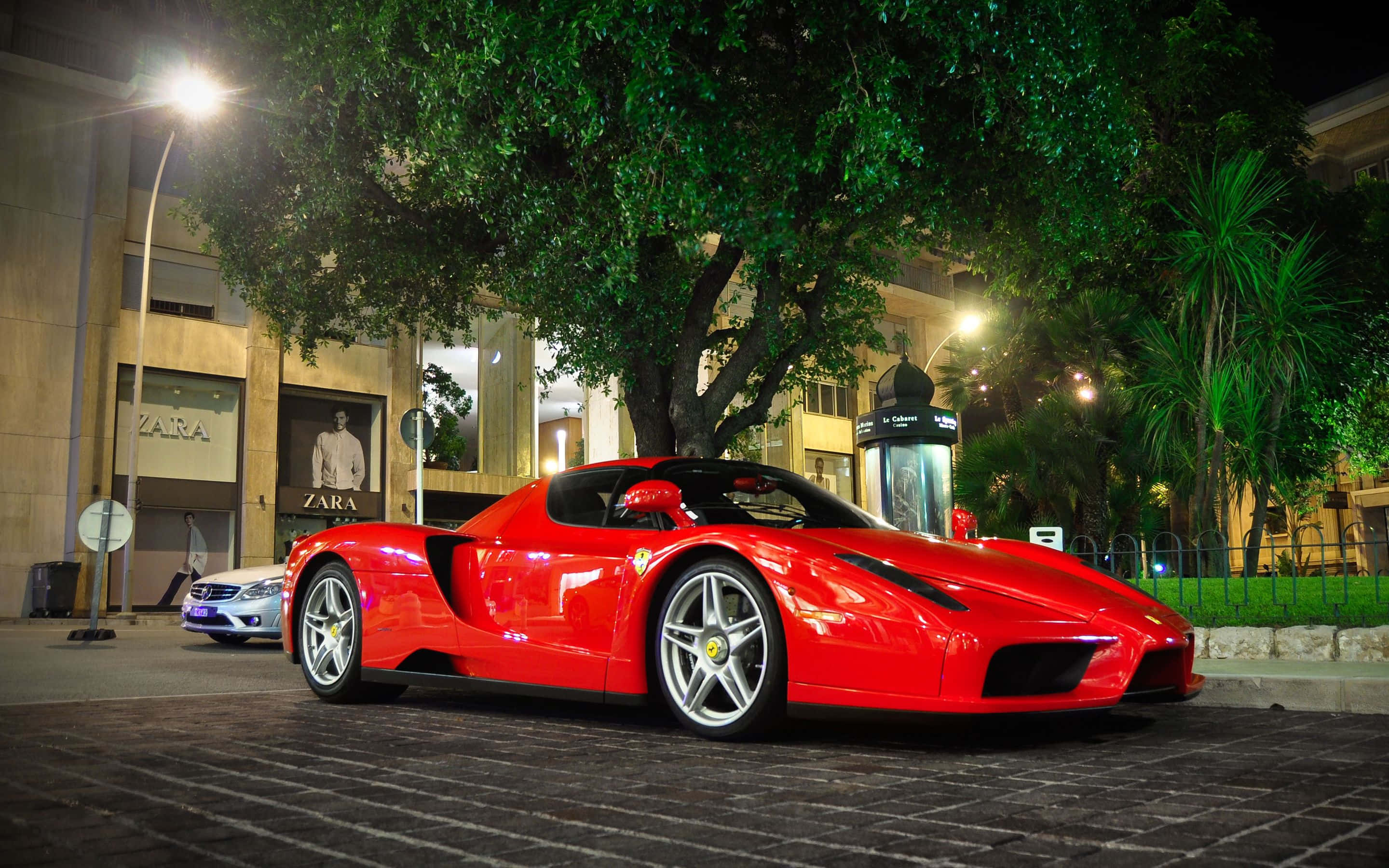 Ferrari Enzo 桌布 壁紙 — 一輛亮麗的紅色法拉利Enzo在夜晚停泊在鵝卵石街道上，位於一棵大樹下，背景是燈火通明的商店門面。